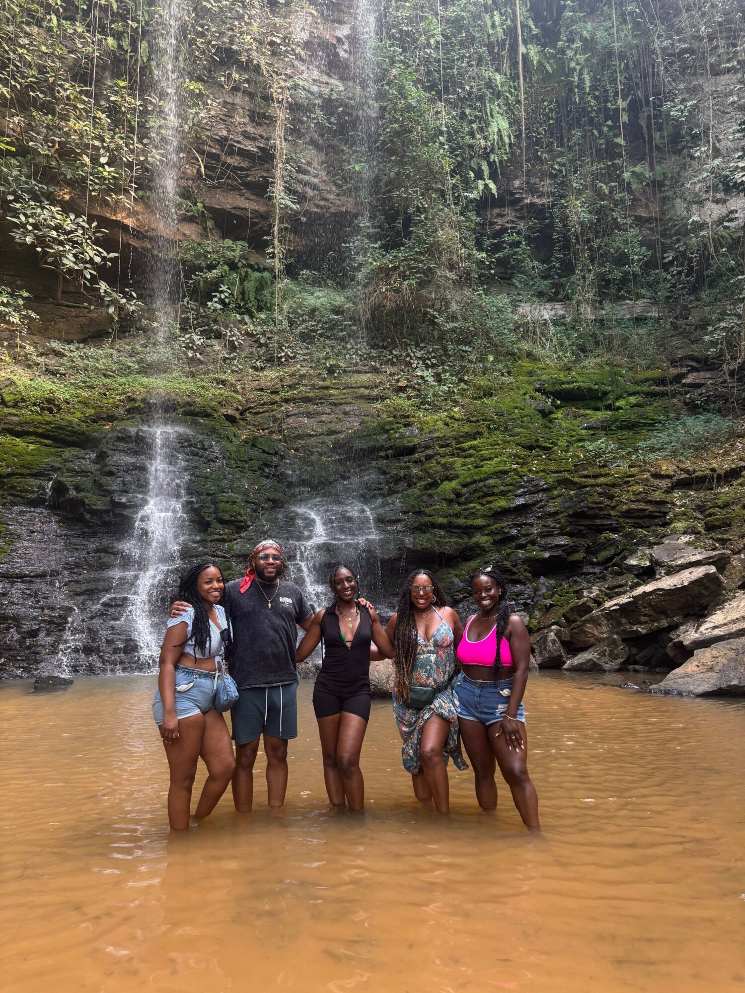 Group of people standing in a shallow river with trees and mossy rocks in the background.