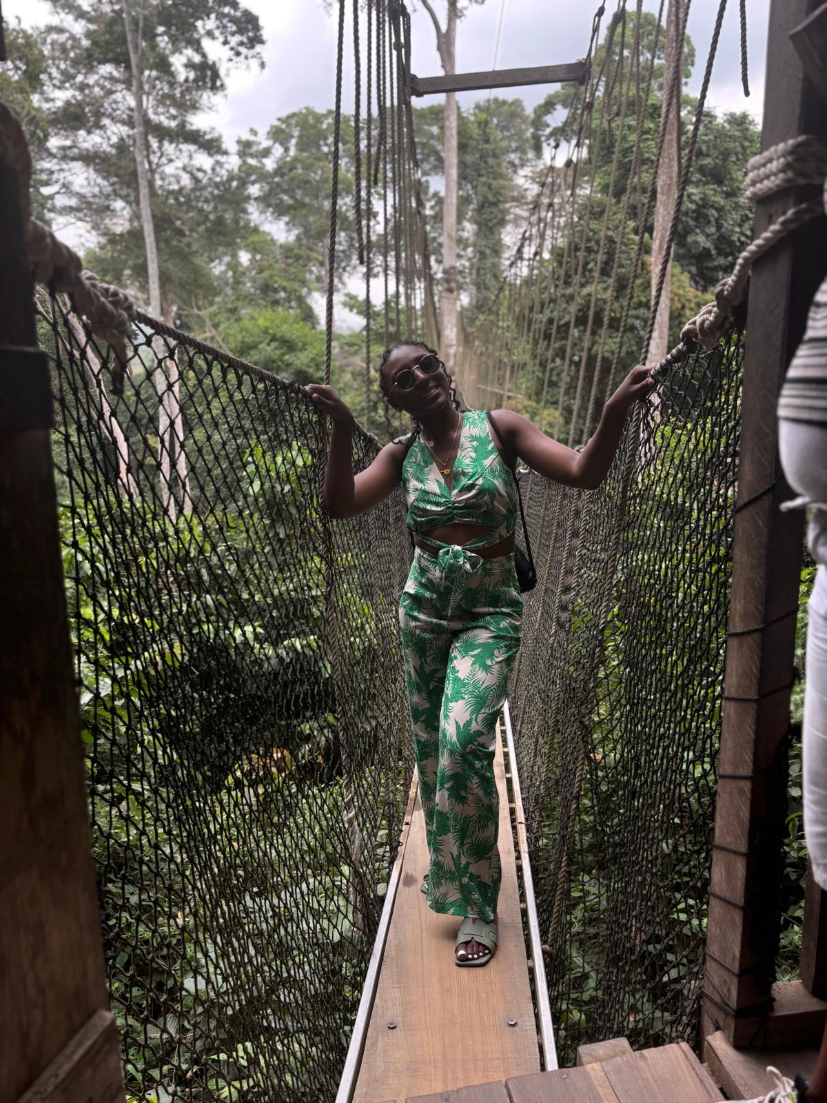Woman in green patterned outfit walking on a narrow suspension bridge in a forest, holding onto the sides.