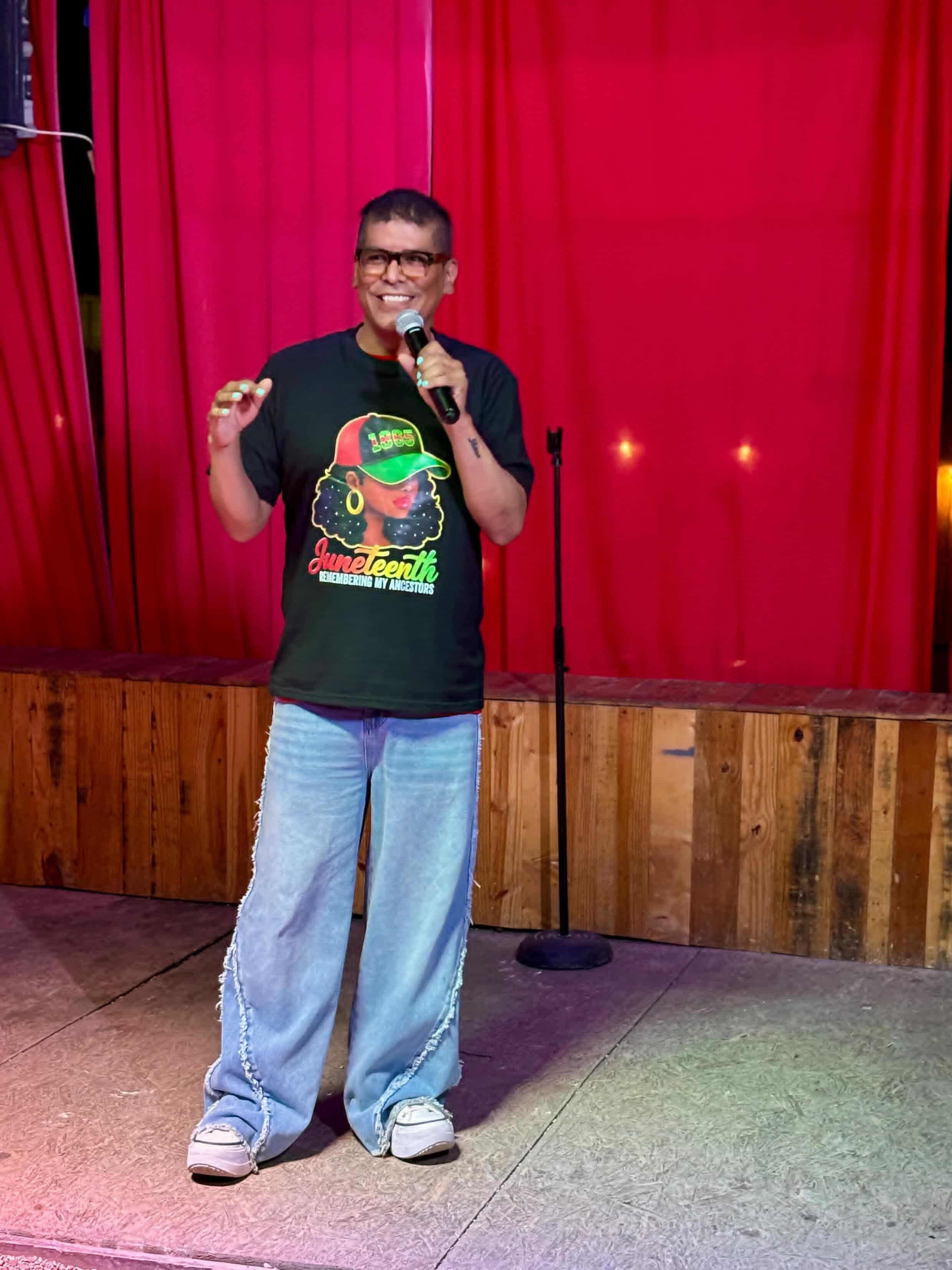 Person standing on stage with red curtains, holding a microphone, wearing glasses, a black T-shirt, and jeans.