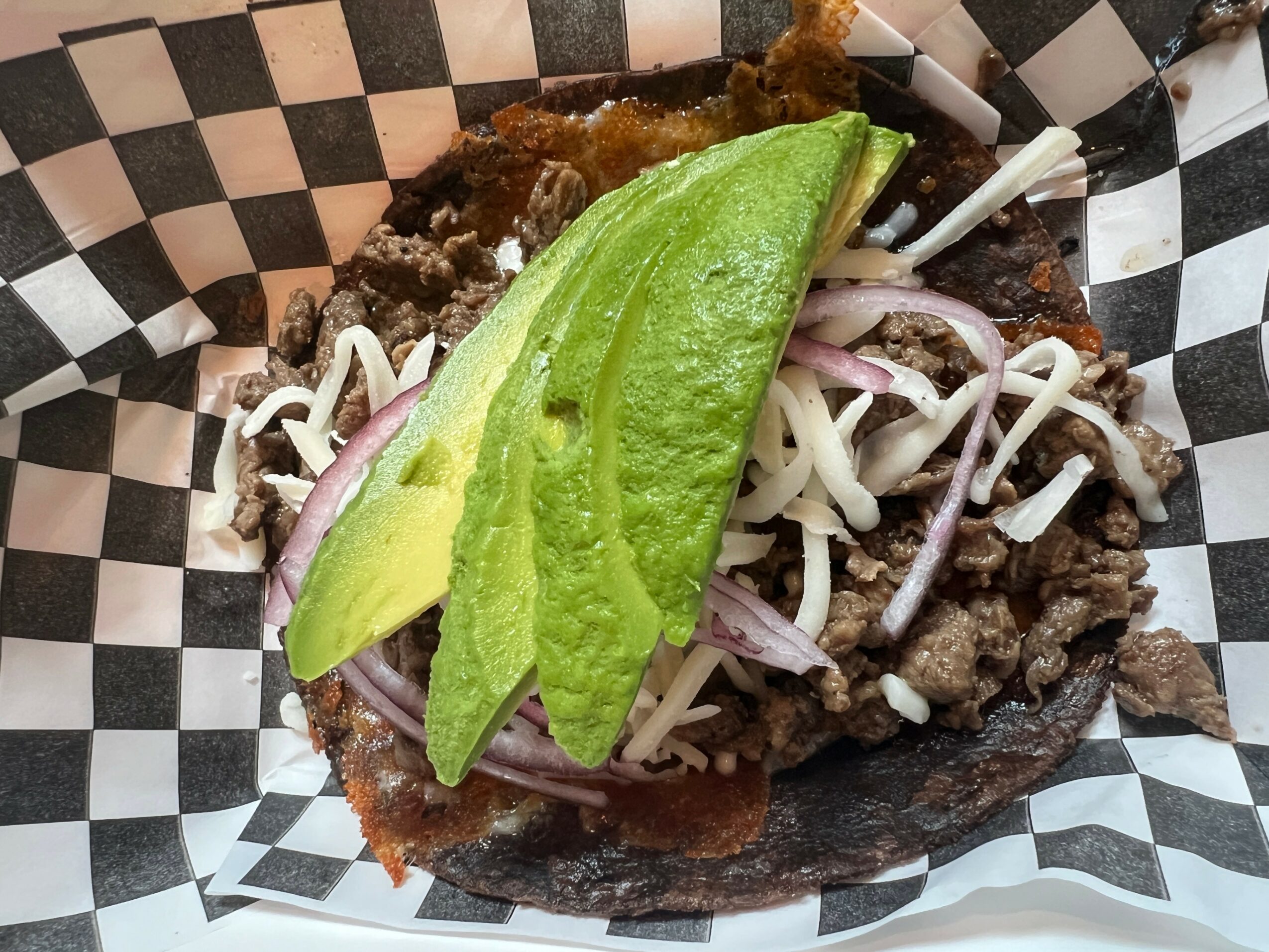 Taco with avocado slices, shredded cheese, onions, and meat in a checkered paper basket.