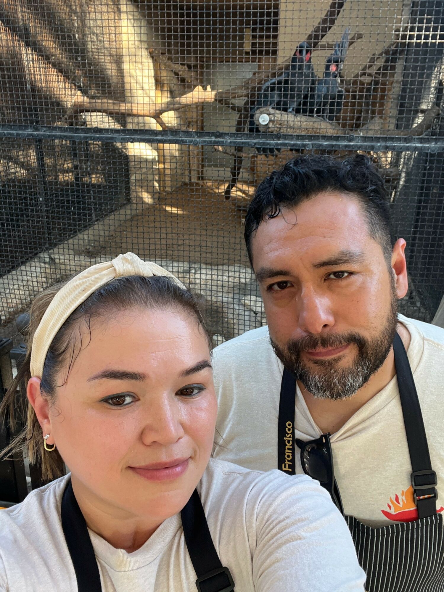 Two people taking a selfie in front of a mesh enclosure with animals inside, smiling at the camera.