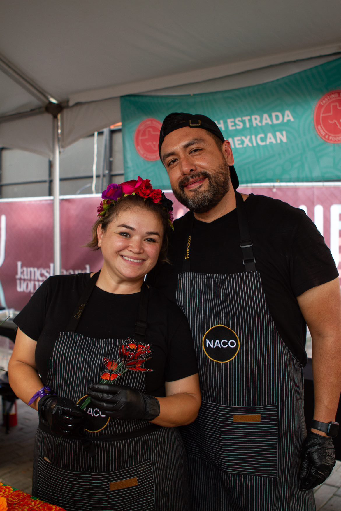 Two people smiling, wearing black aprons, standing in front of a teal banner with red and white text, inside a tent.