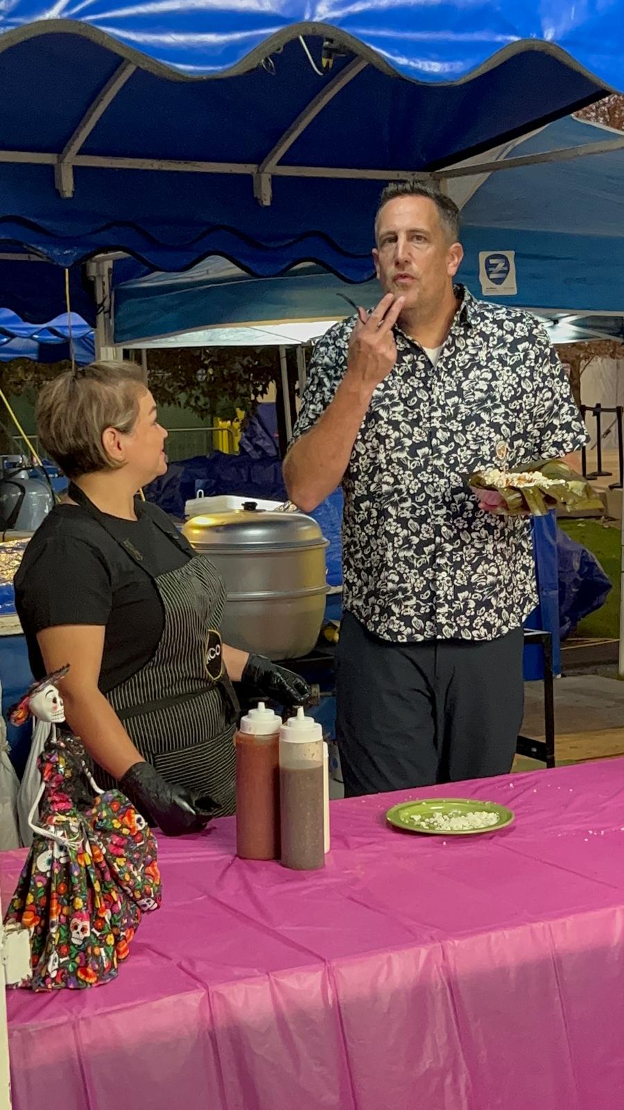 Two people at a table with condiments, under a blue canopy, with a man holding a plate and a woman nearby.