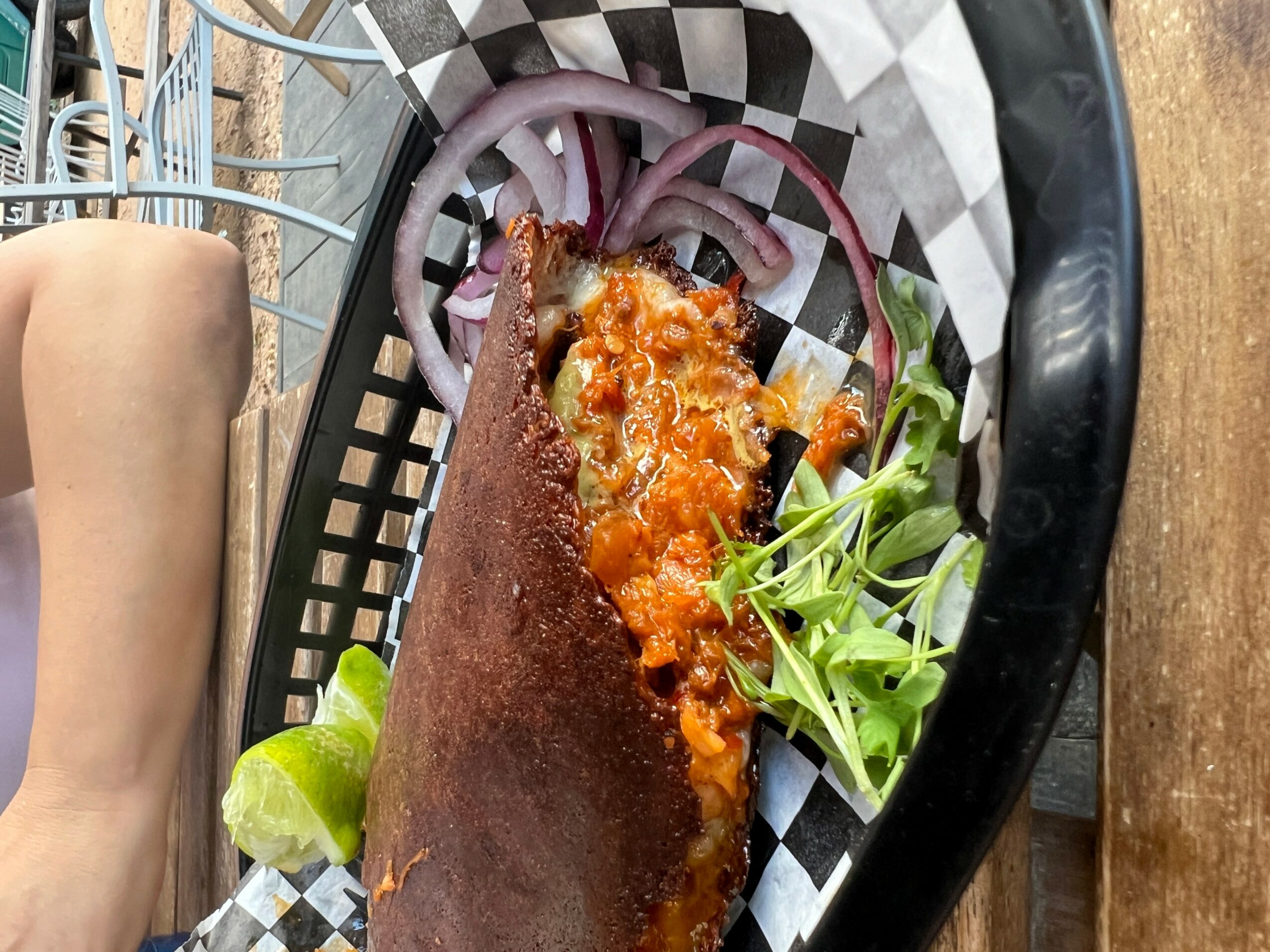 Fried fish fillet with sauce, onion slices, and greens on checkered paper in a basket, with a lime wedge nearby.
