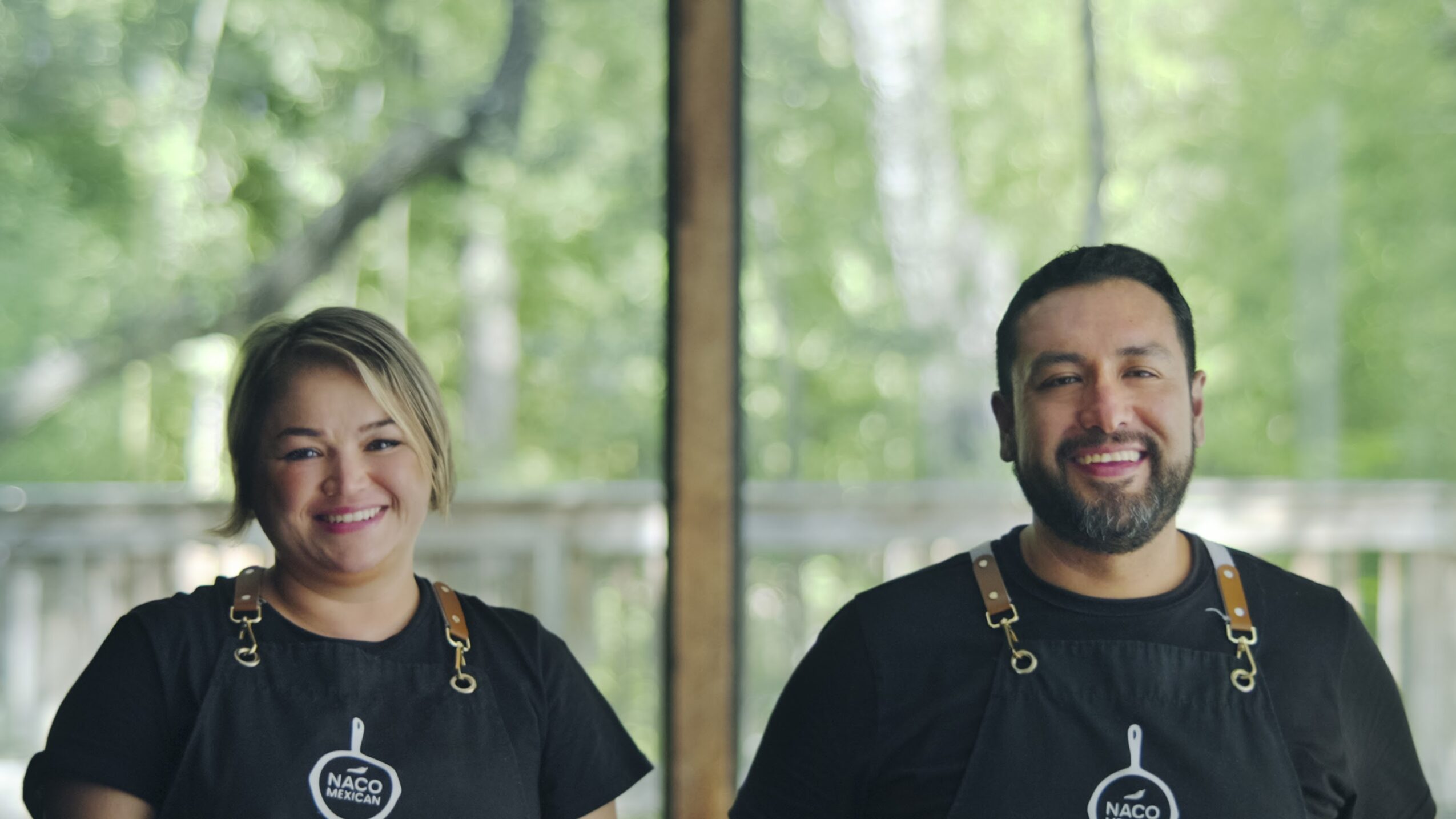 Two smiling people wearing black aprons stand indoors with a green, leafy background visible through windows.