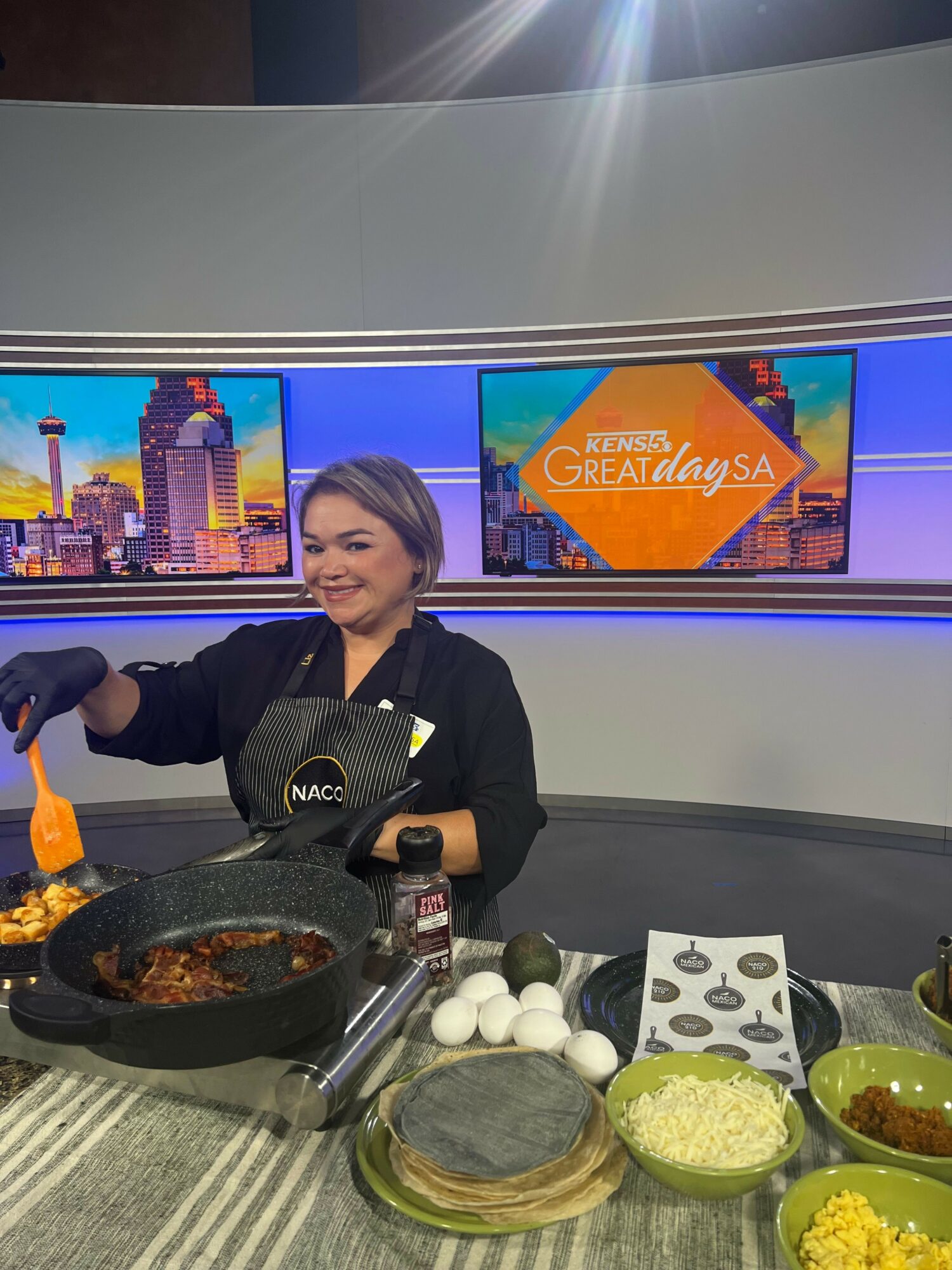 Woman cooking at a kitchen counter with bowls of ingredients, TV screens displaying a cityscape, and a gray countertop.