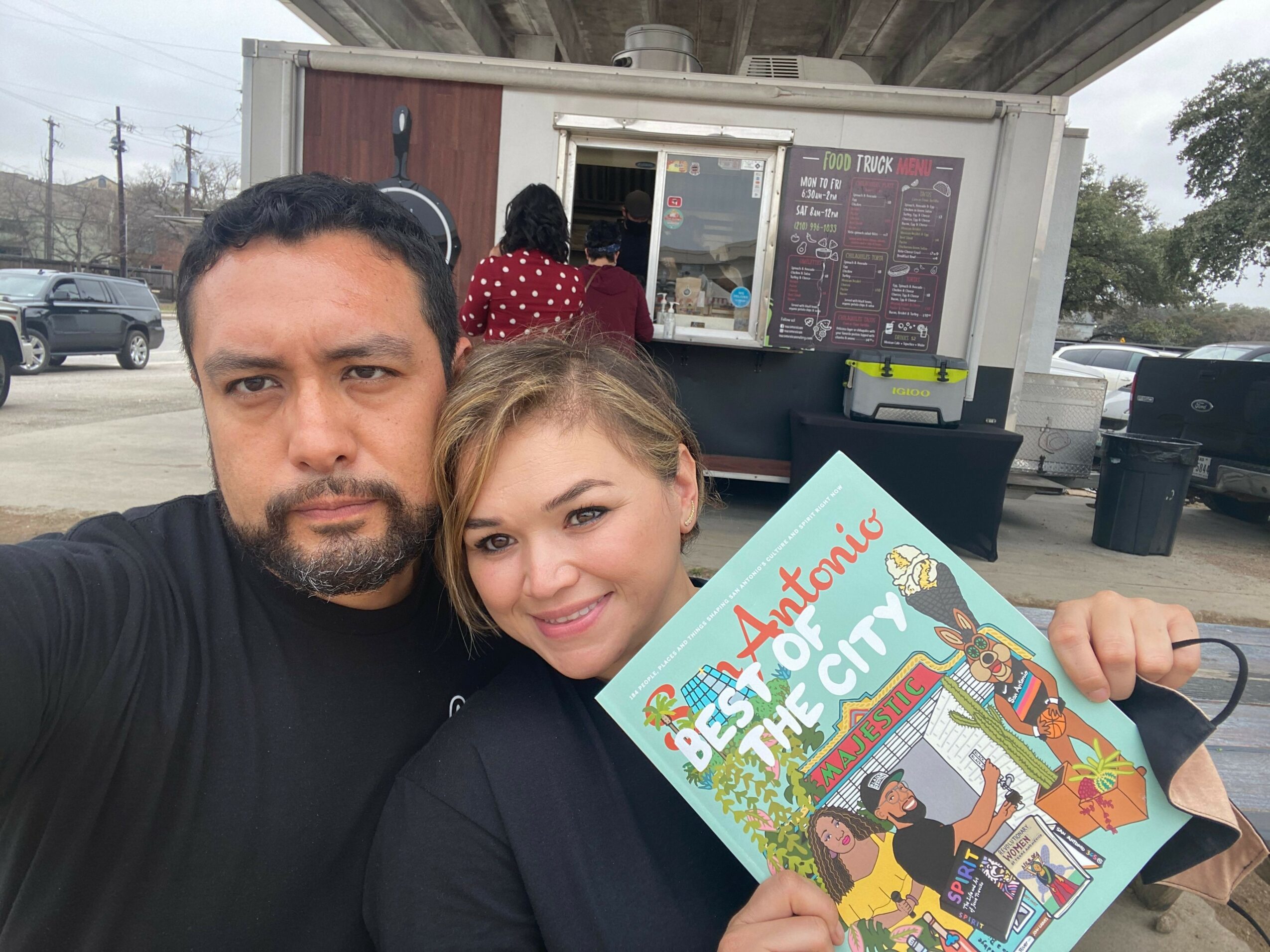 Two people, a woman and a man, pose outdoors with a book, smiling at the camera, with a building and trees in the background.