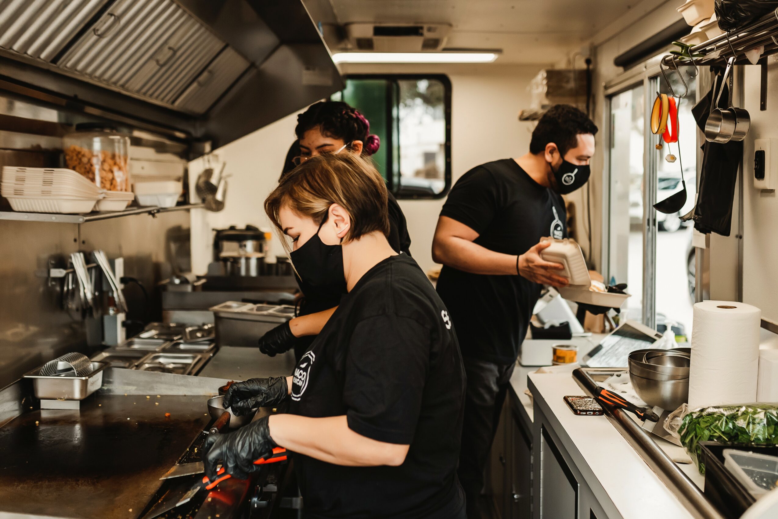 Three people working in a kitchen, wearing black masks, preparing food at a counter with cooking equipment and utensils.