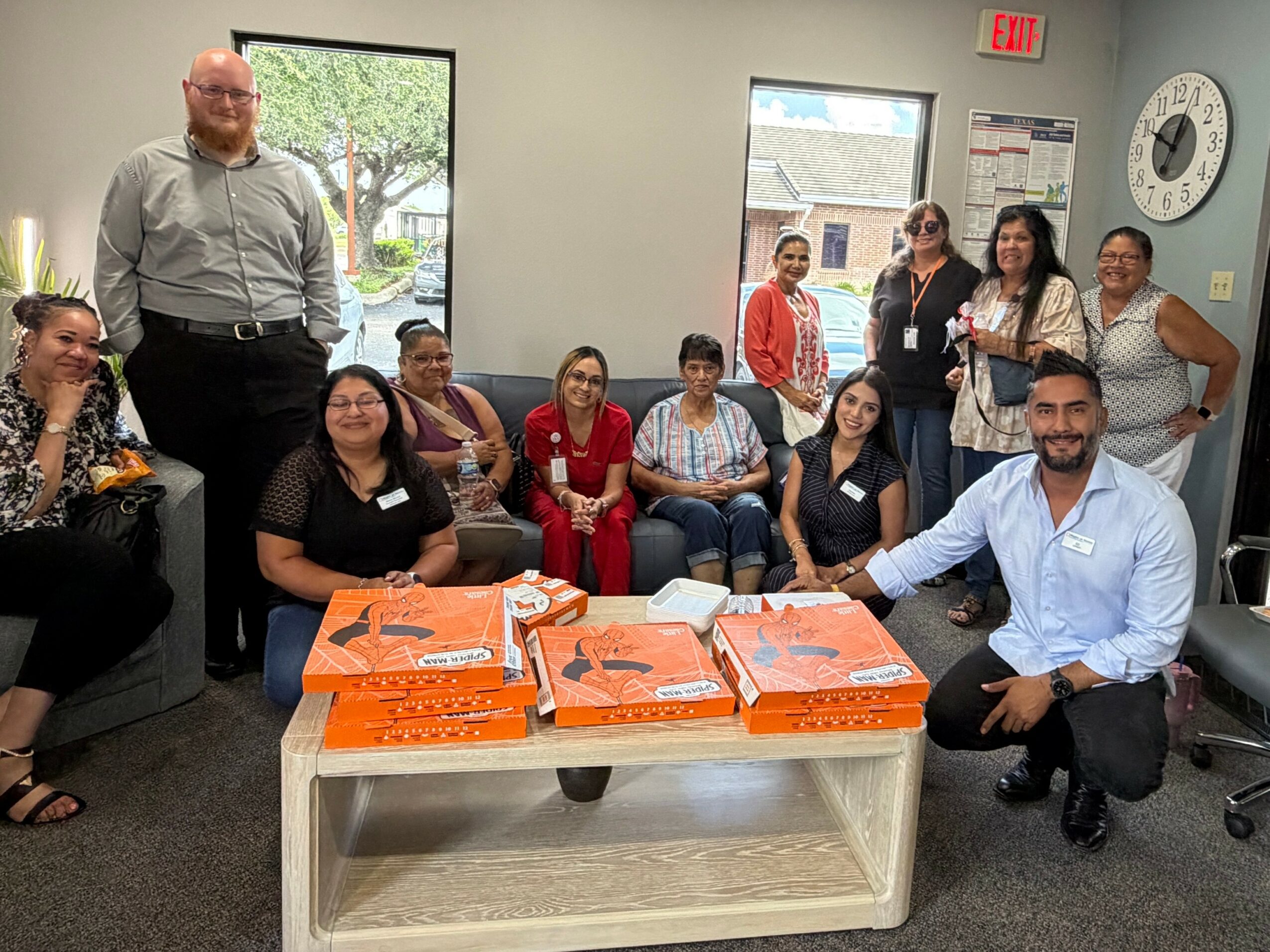 Group of people gathered in a room with a table holding orange boxes, some seated and others standing, smiling.