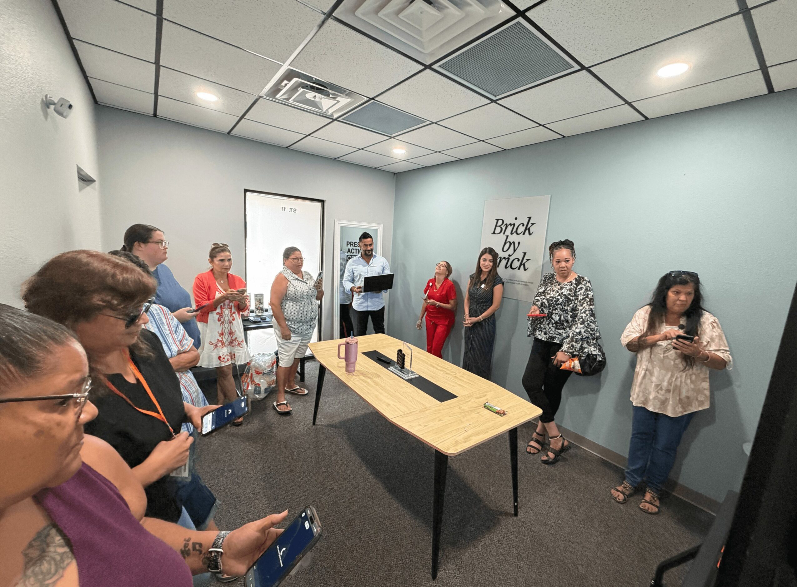 Group of people standing in a room with a table, some holding phones, and a wall with a sign that says 'Brick by brick'.