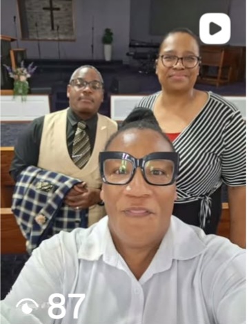 Three people posing indoors, one in front wearing glasses and a white shirt, two behind, one in a vest and tie, the other in a striped shirt.