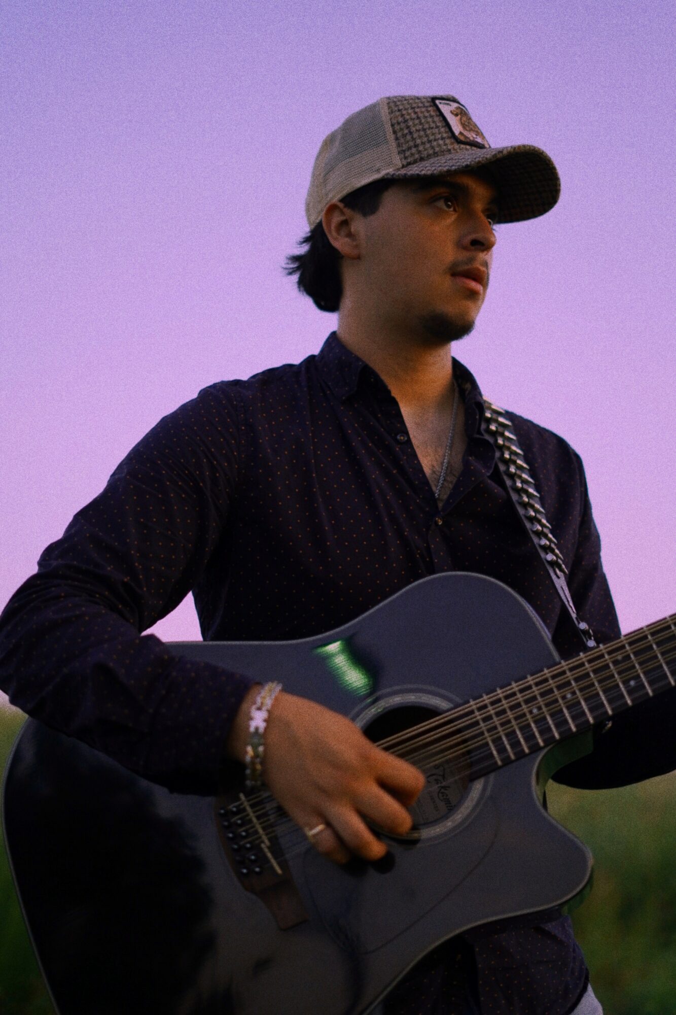 Young man wearing a cap playing an acoustic guitar outdoors during sunset or dusk.