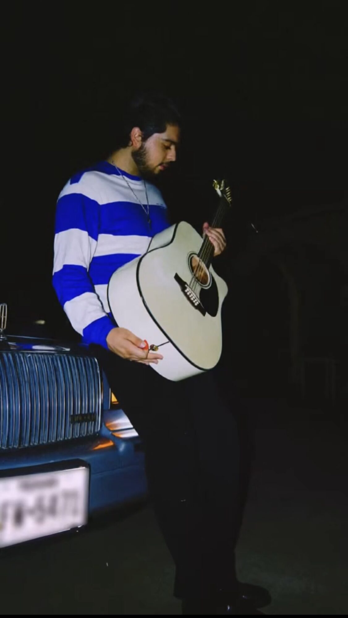 Person with dark hair and beard playing an acoustic guitar outdoors at night, wearing a blue and white striped sweater.