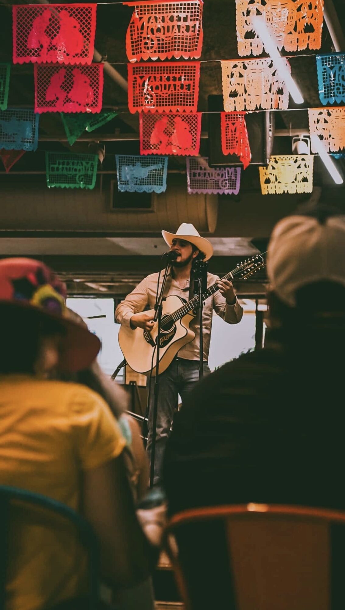 Person wearing a hat playing guitar on stage with colorful papel picado banners overhead, audience in foreground.