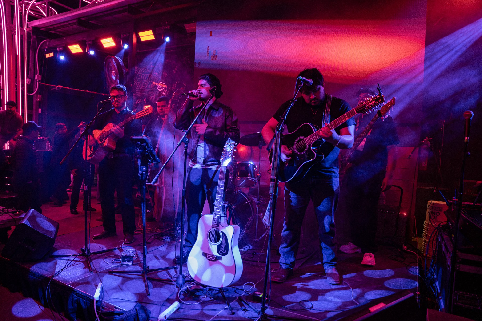 Band performing on stage with colorful lighting, three musicians playing guitars and singing, with a drum set in the background.