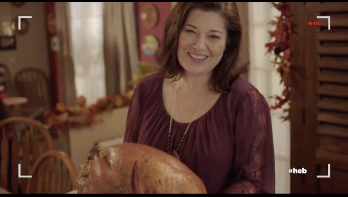 Woman smiling in a decorated room holding a large cooked turkey, autumn-themed decorations visible in the background.