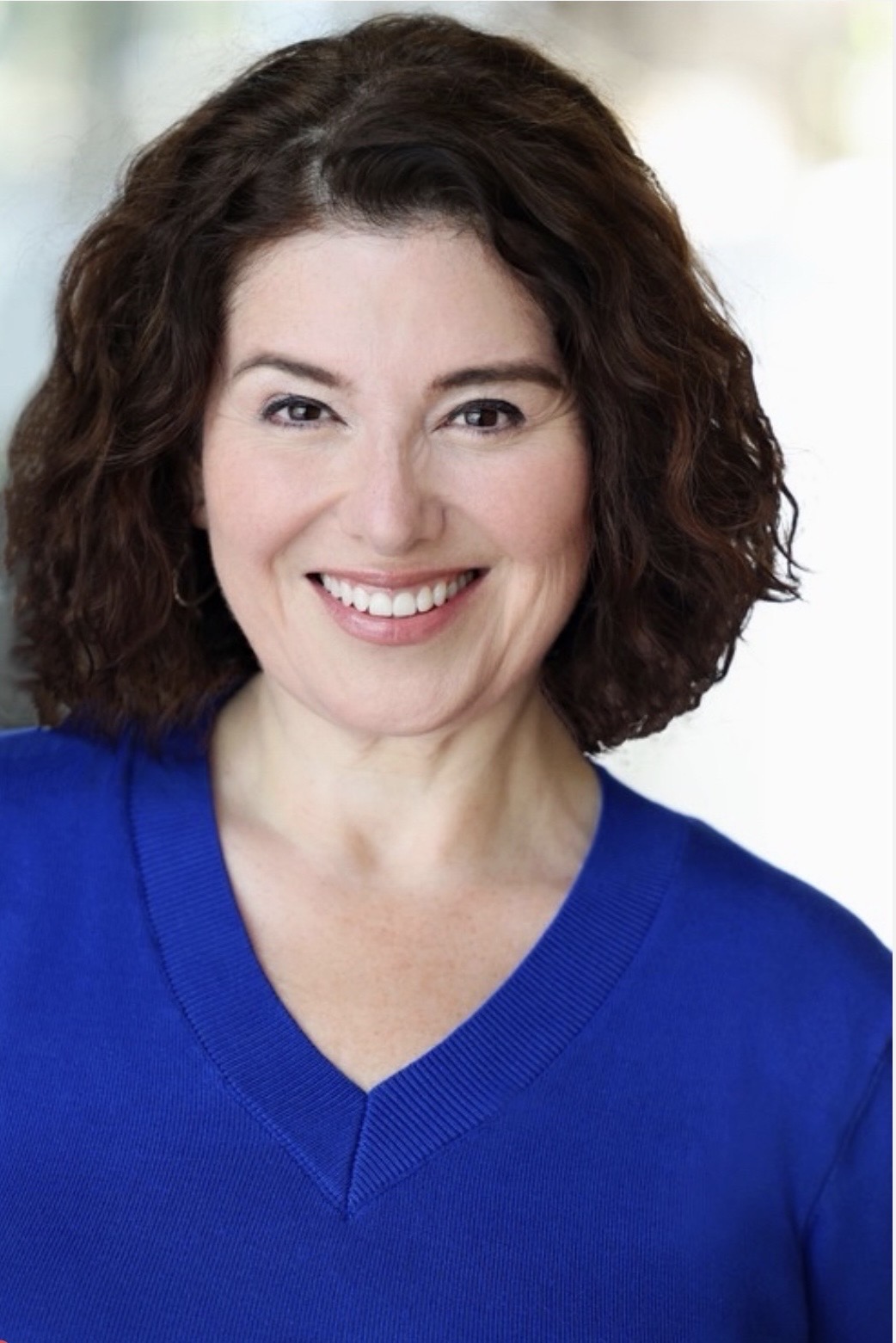 Smiling woman with curly dark hair wearing a blue top, outdoors with blurred background.