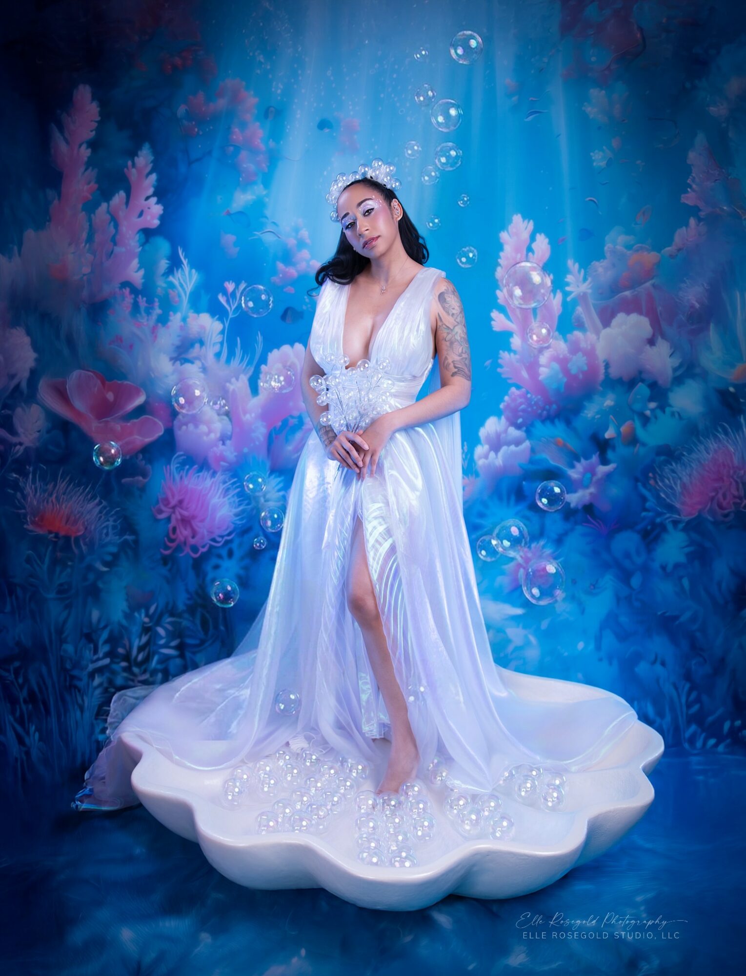 Woman in a white dress standing on a shell-shaped platform underwater scene with coral and bubbles, wearing a tiara.