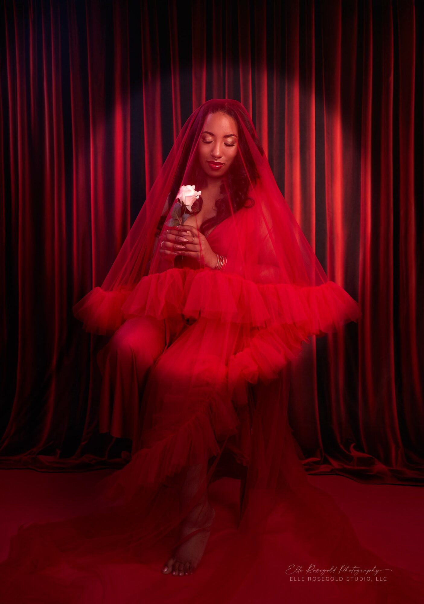 Woman in red dress with veil holding a white flower, standing in front of red curtains.