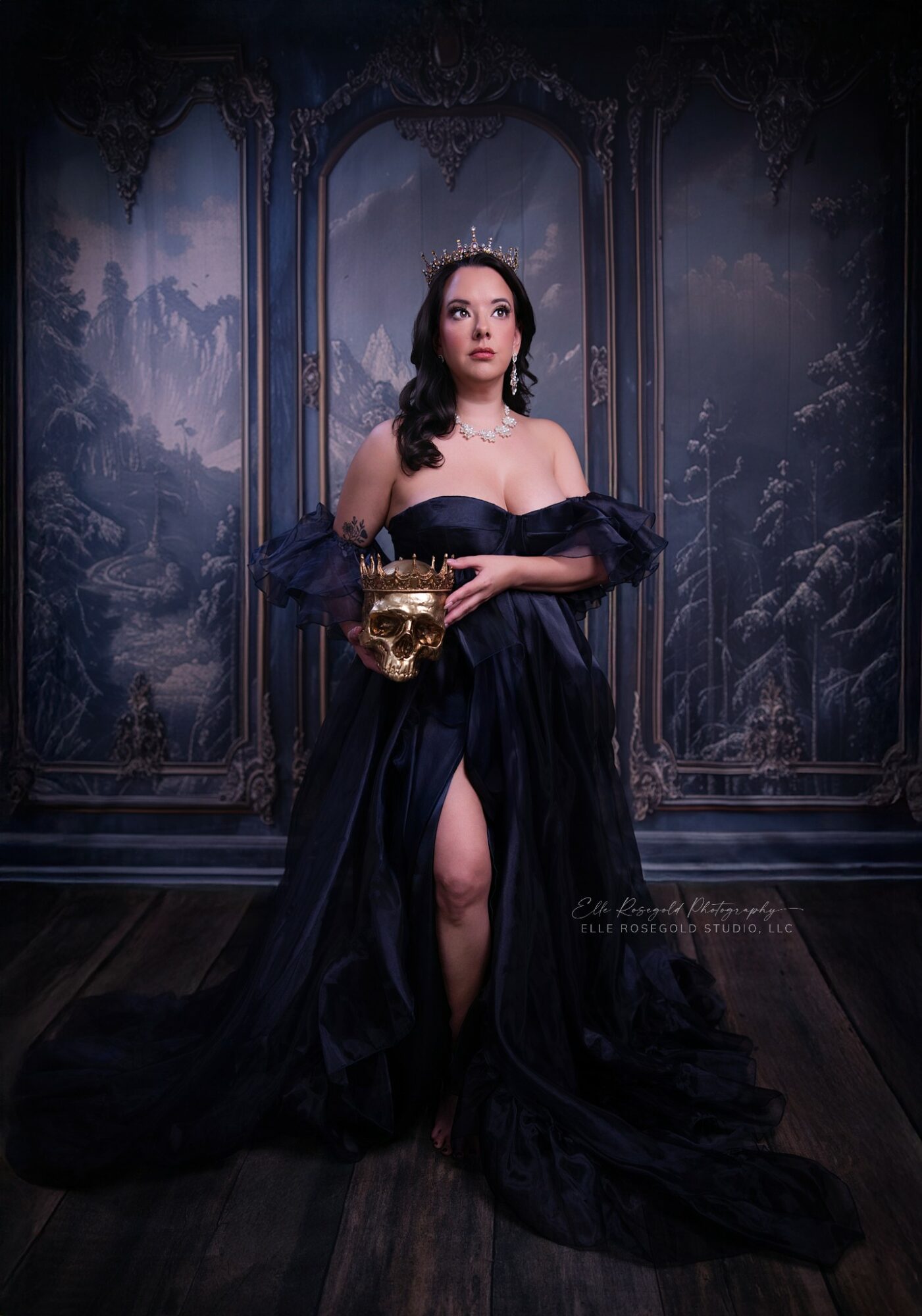 Woman in black gown holding a gold skull, standing in a dark, ornate room with painted walls.