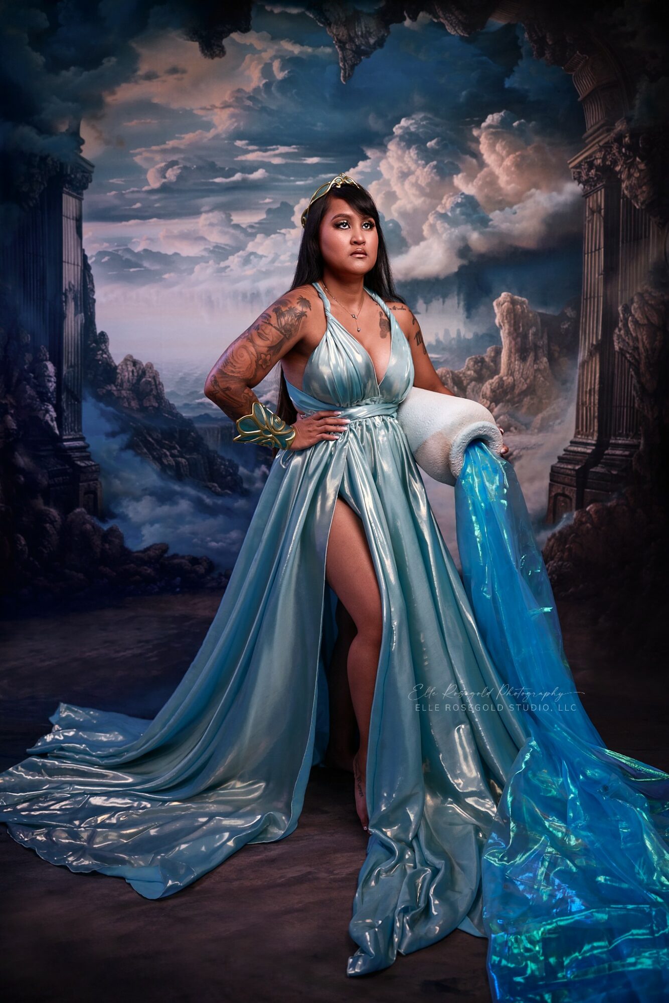 Woman in a flowing blue gown with tattoos, standing in a fantasy landscape with mountains and cloudy sky, holding a white and blue fabric.