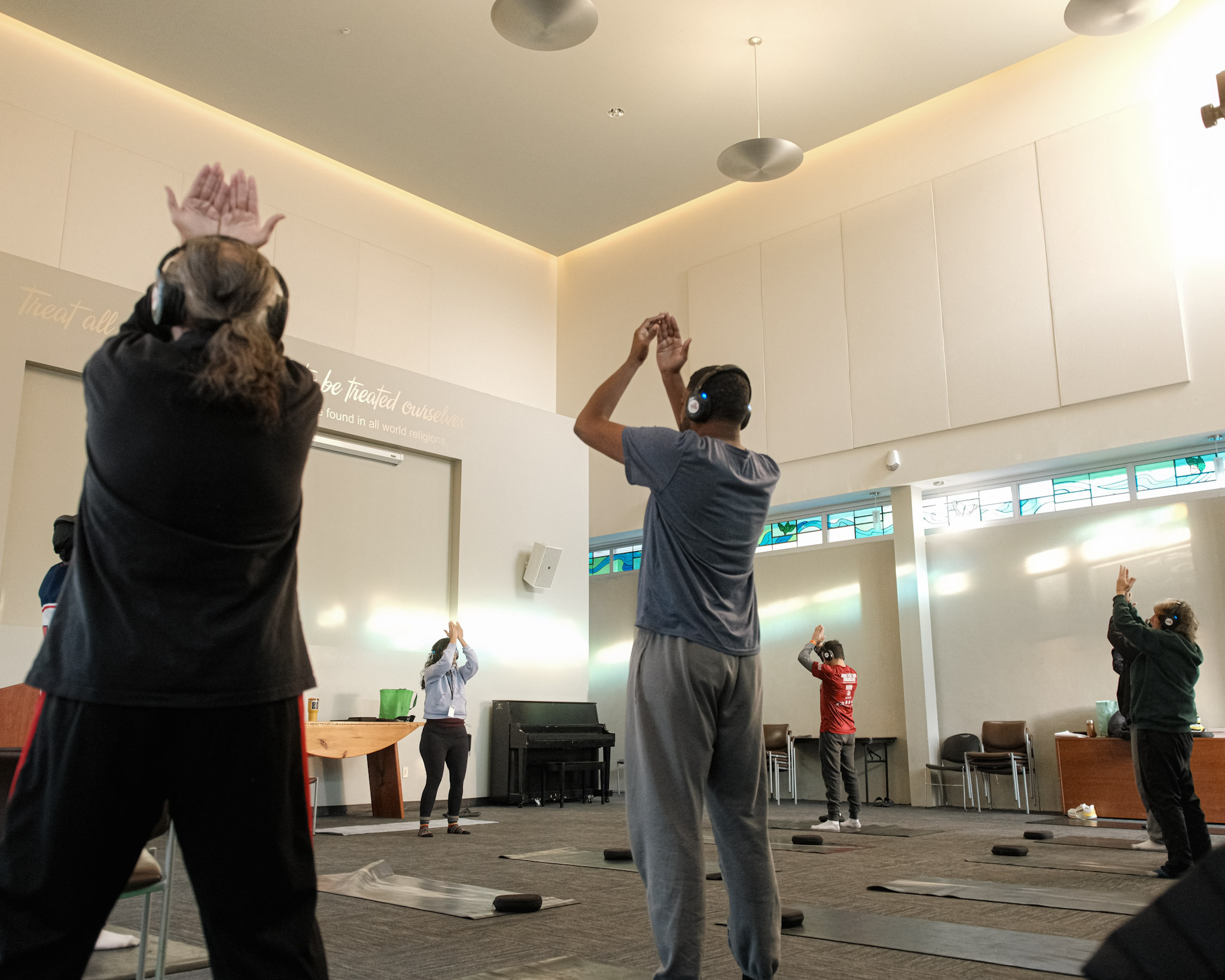 People standing in a spacious room, some raising their hands, with natural light and a piano in the background.