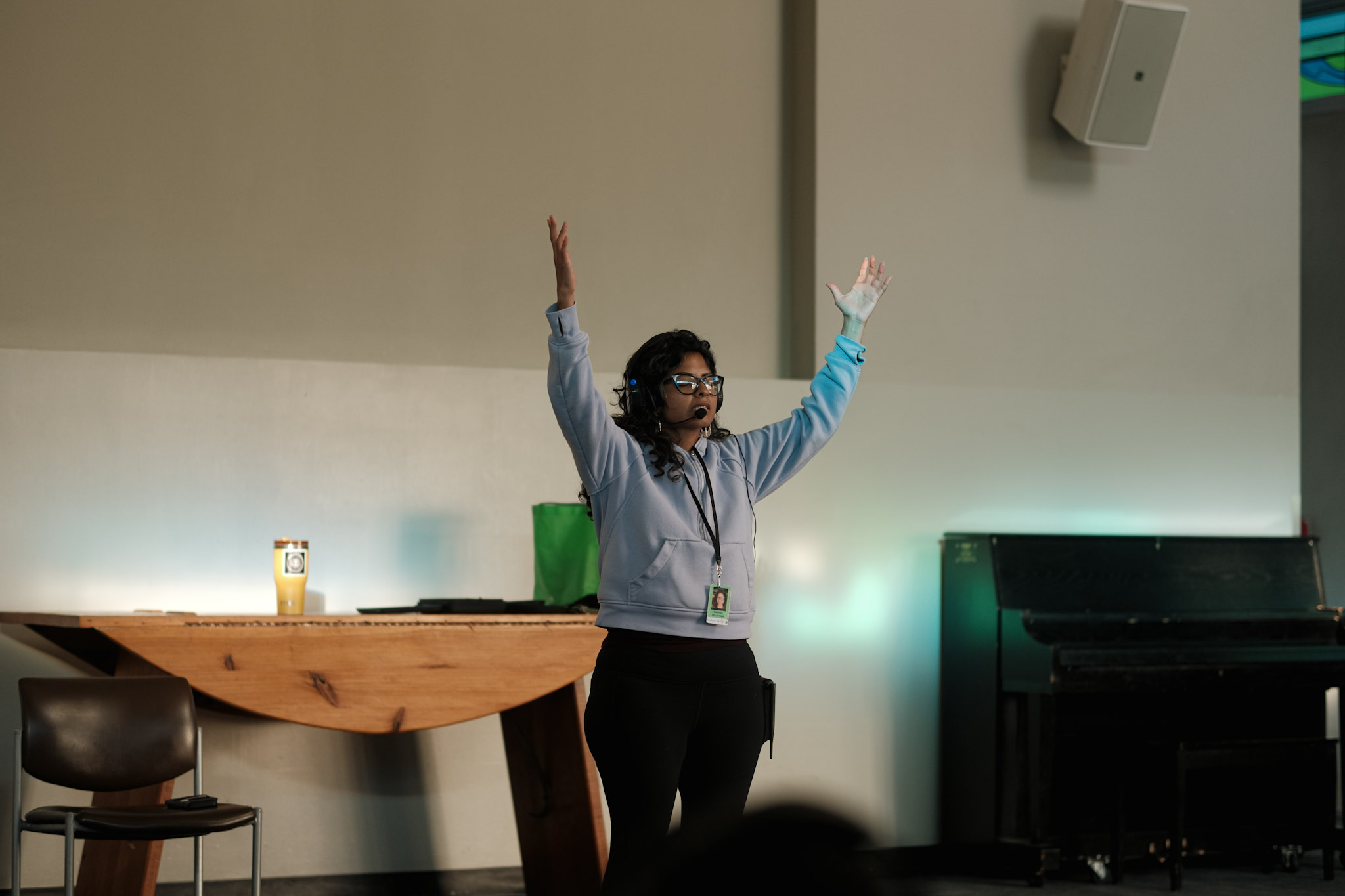 Person with glasses and curly hair raises both arms, standing in a room with a piano and a wooden table.