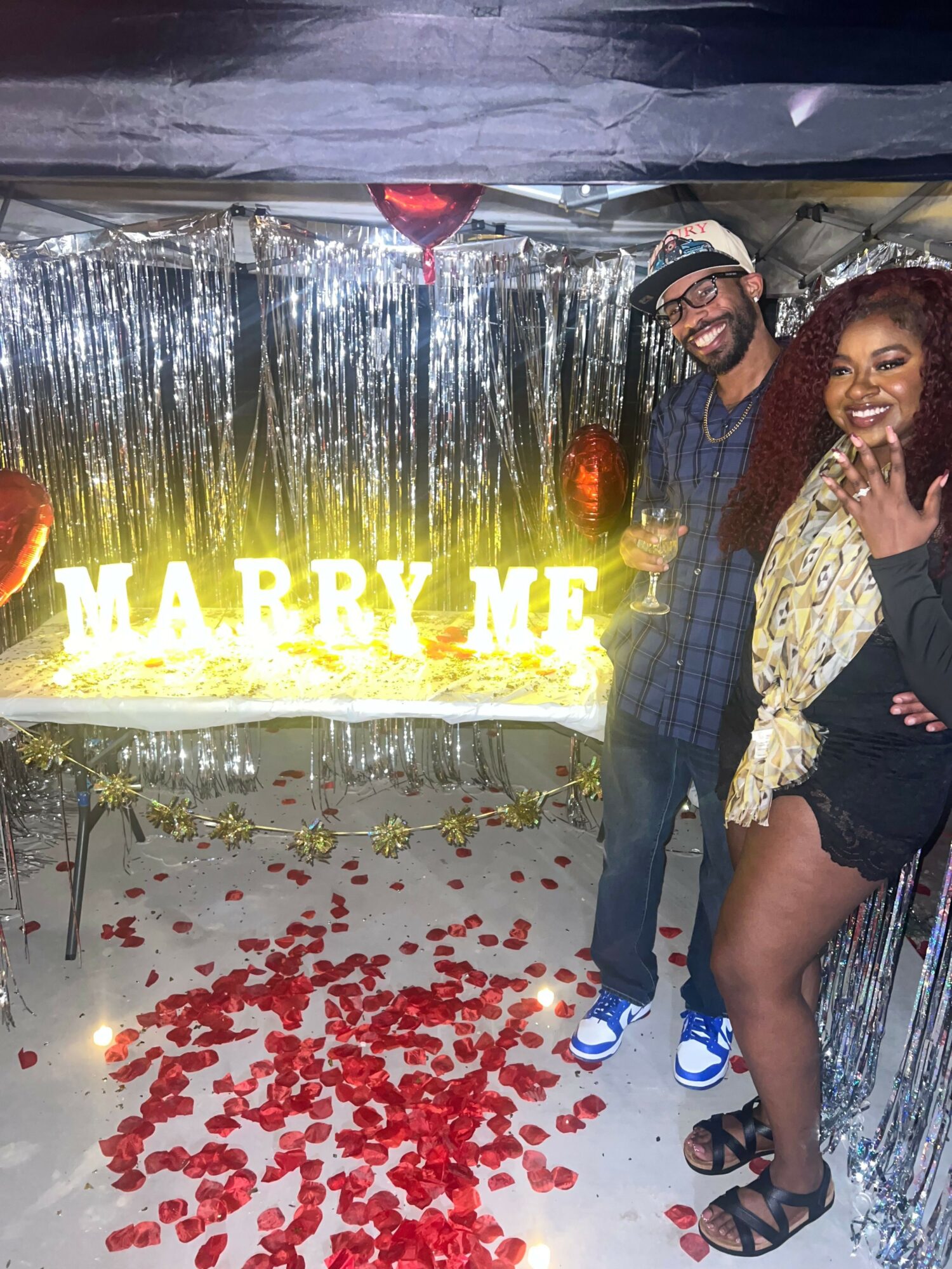 Two people posing with a decorated backdrop and a large illuminated sign that says 'WARRRY ME'. Red rose petals are scattered on the floor.