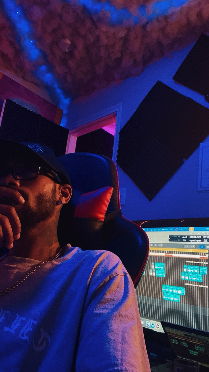 Person wearing glasses and a cap in a music studio with soundproof panels and a computer screen.