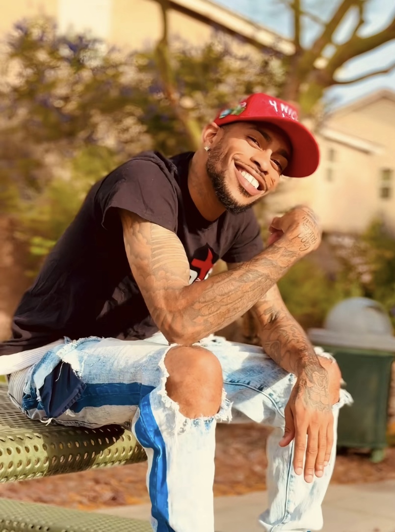 Smiling man with tattoos wearing a red cap and ripped jeans, sitting outdoors with trees and buildings in background.