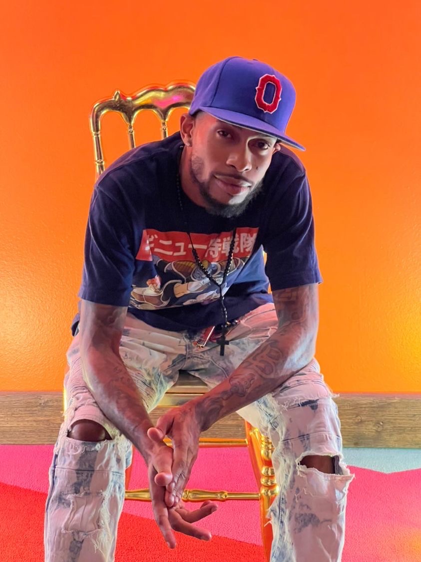Man sitting on a gold chair with an orange background, wearing a blue cap and casual clothes, looking at the camera.