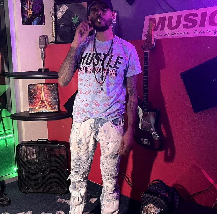 Man with tattoos and dreadlocks standing in a music studio, holding a cigarette, wearing a white T-shirt and light-colored pants.