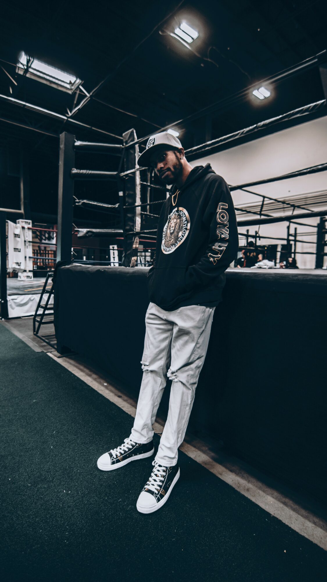 Person standing in a boxing ring, wearing a hoodie, cap, and sneakers, with boxing equipment in the background.