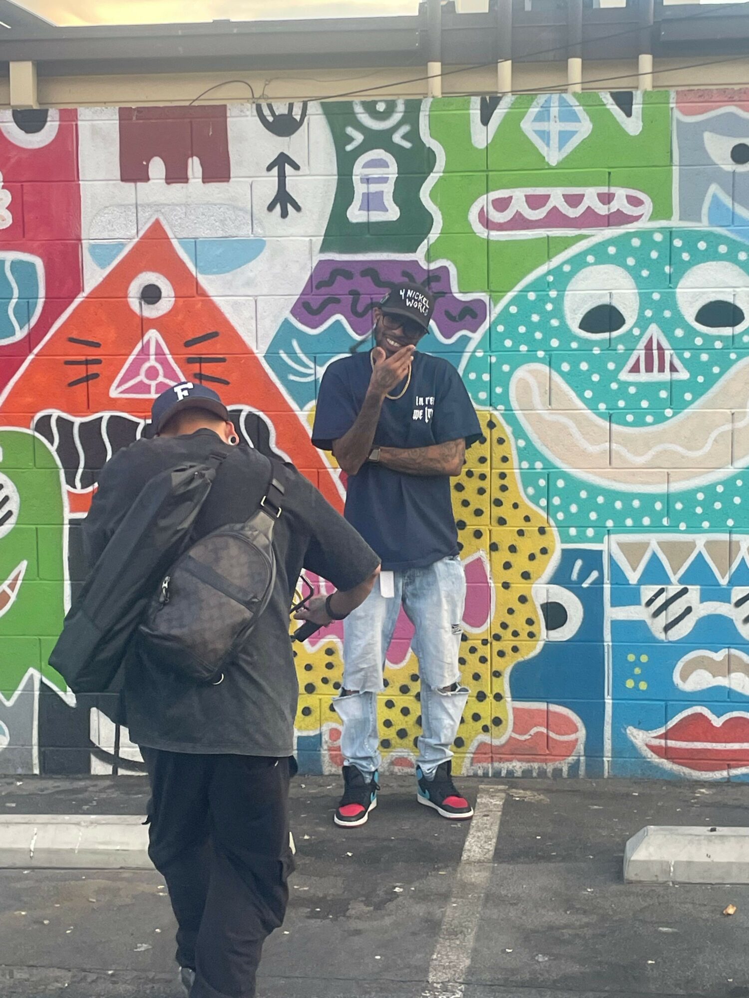 Two people stand in front of a colorful mural with abstract shapes and animals, one taking a photo, the other looking at the mural.