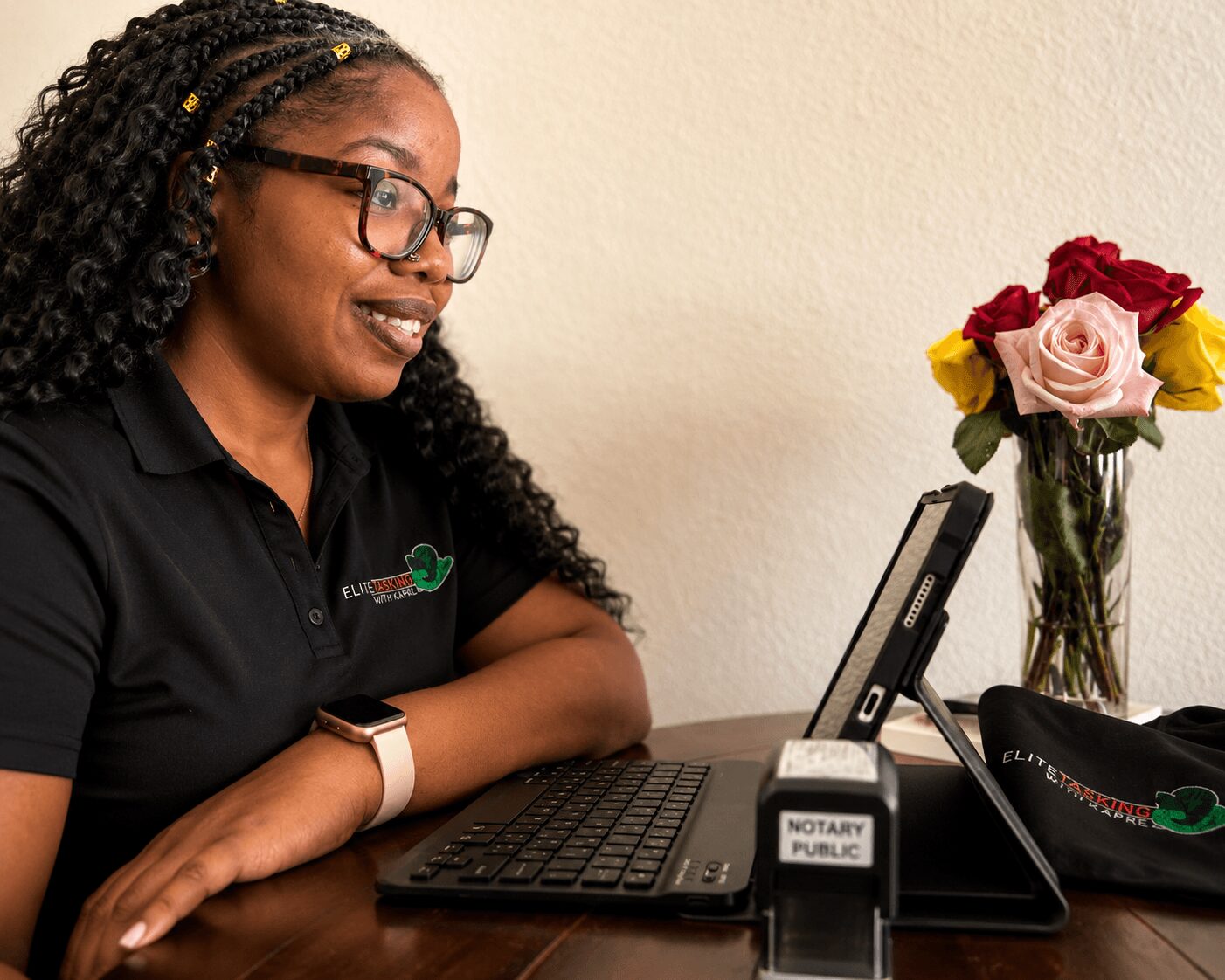 Woman with glasses and curly hair using a laptop at a desk with a vase of flowers nearby.