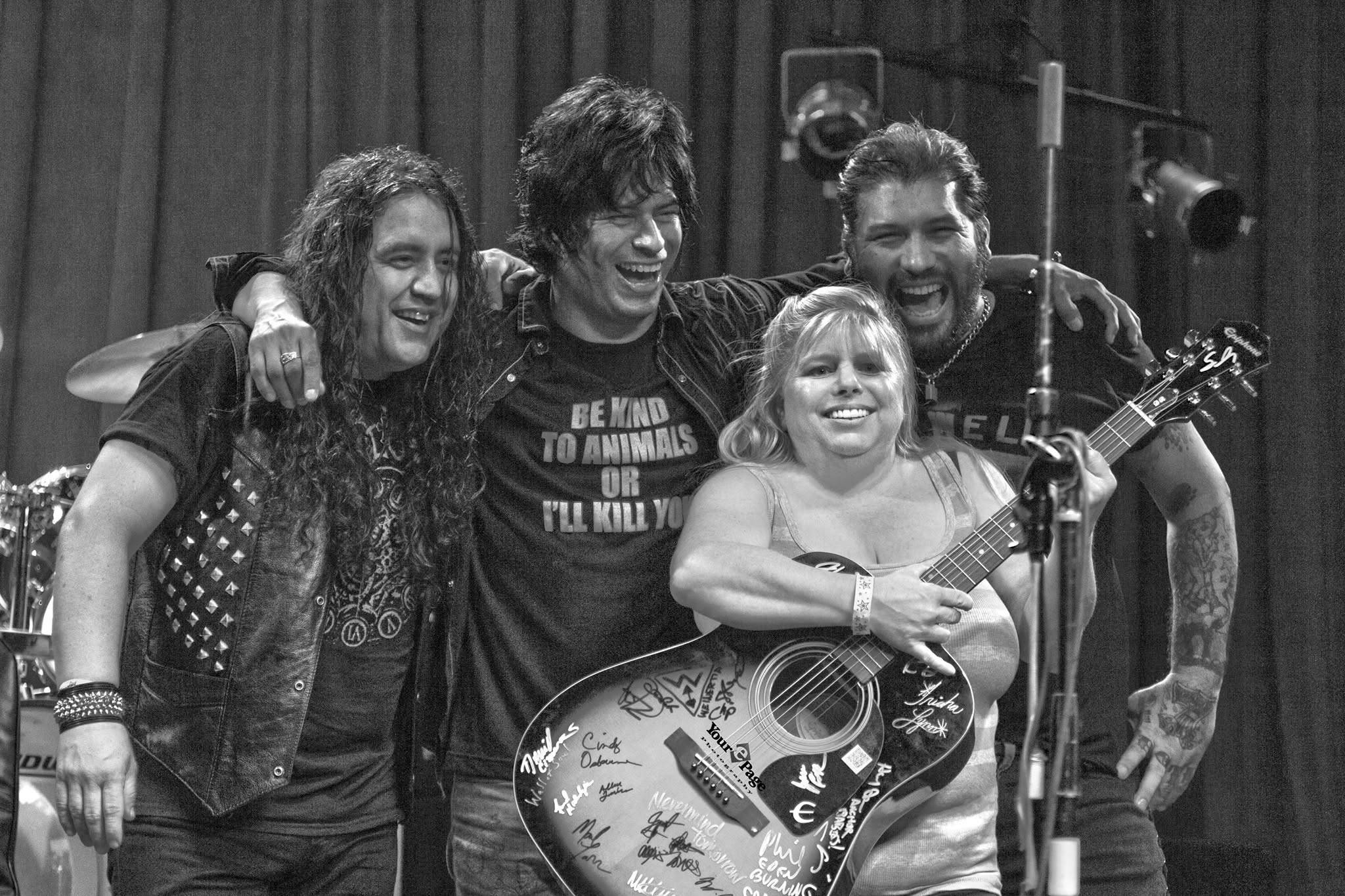 Four people, three adults and one child, smiling and posing with a guitar on stage, black and white photo.