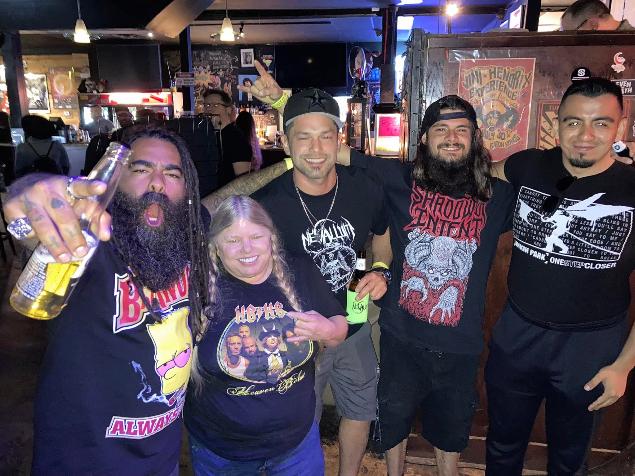 Group of five people smiling in a bar, some holding drinks, with colorful posters and bar lights in background.