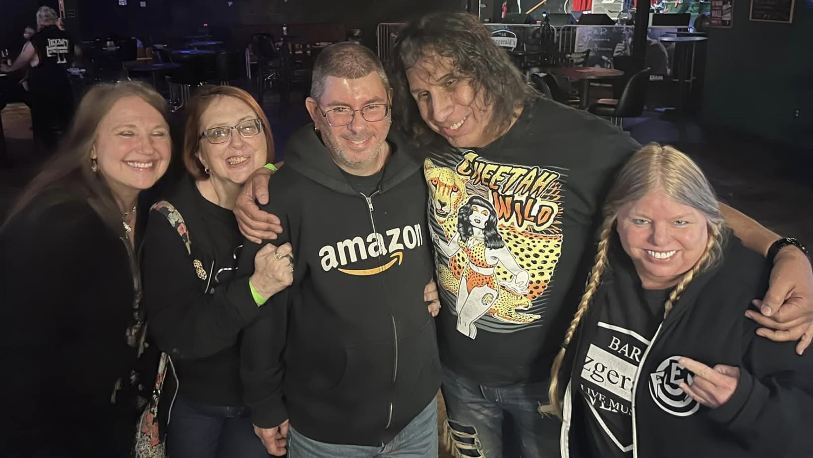 Group of five people smiling and hugging in a dimly lit indoor setting.