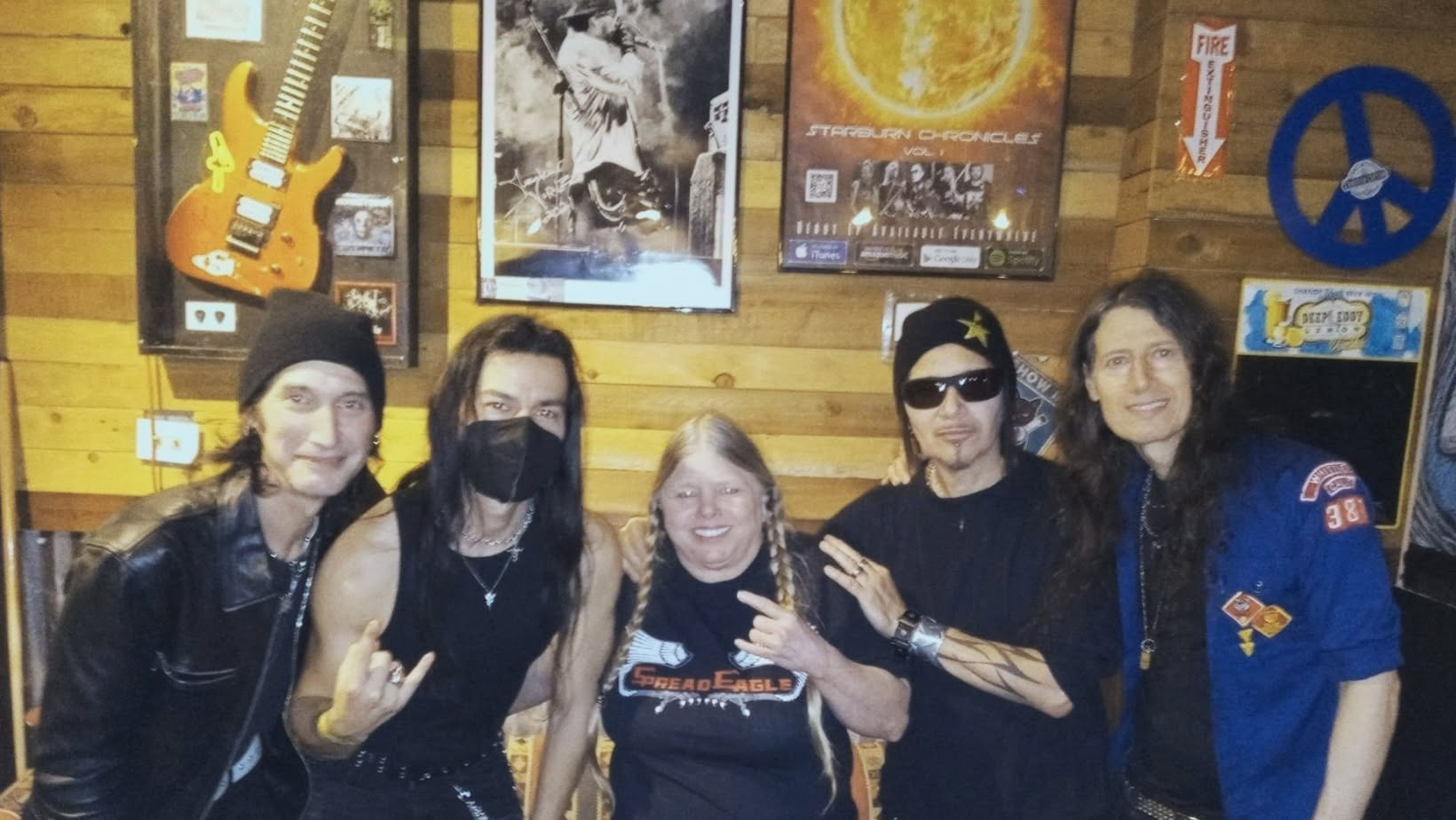 Group of five people standing indoors, some wearing black masks and sunglasses, with posters and decorations on the wall behind them.