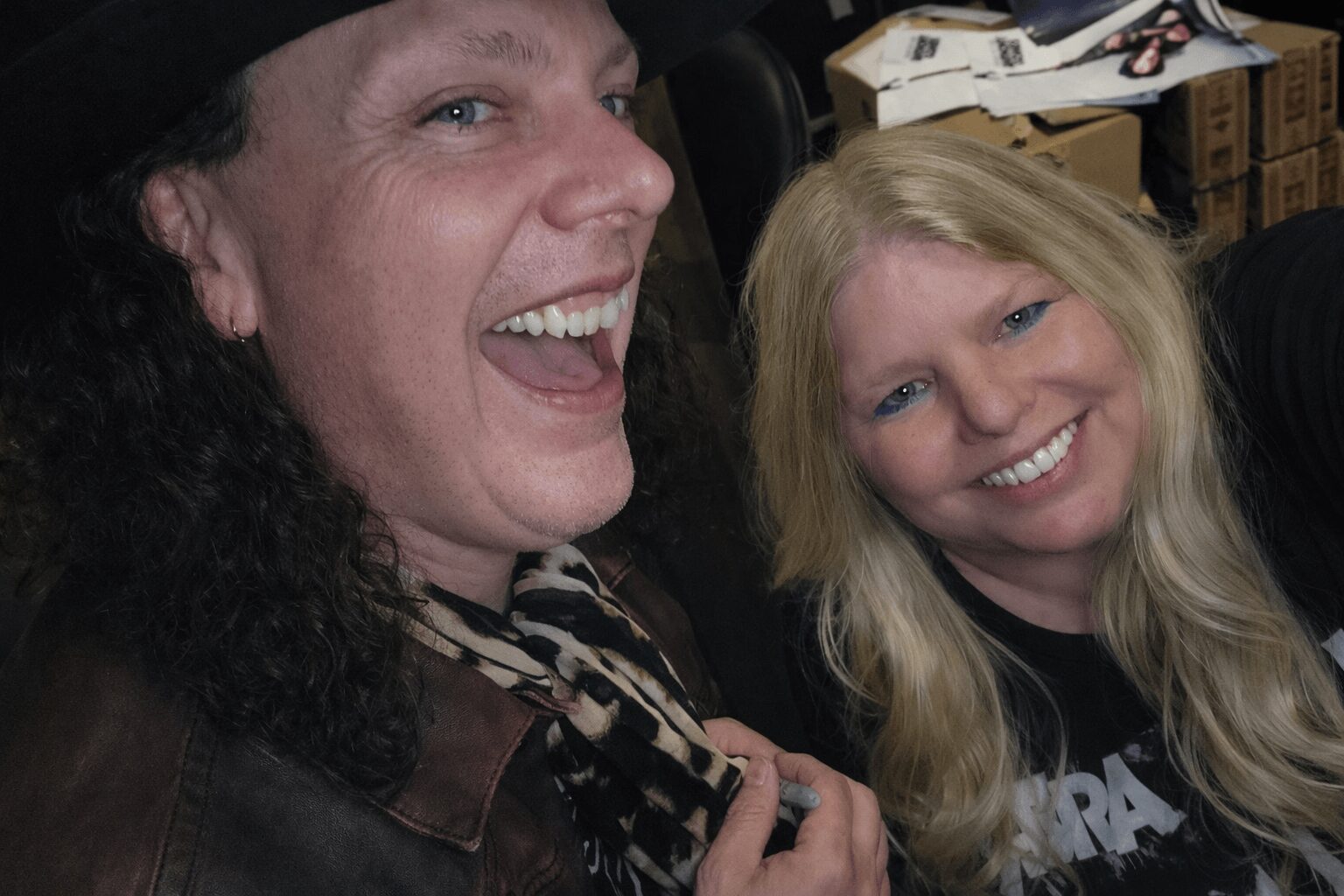 Two smiling people taking a selfie indoors, with a cluttered background. One has dark curly hair, the other has long blonde hair.