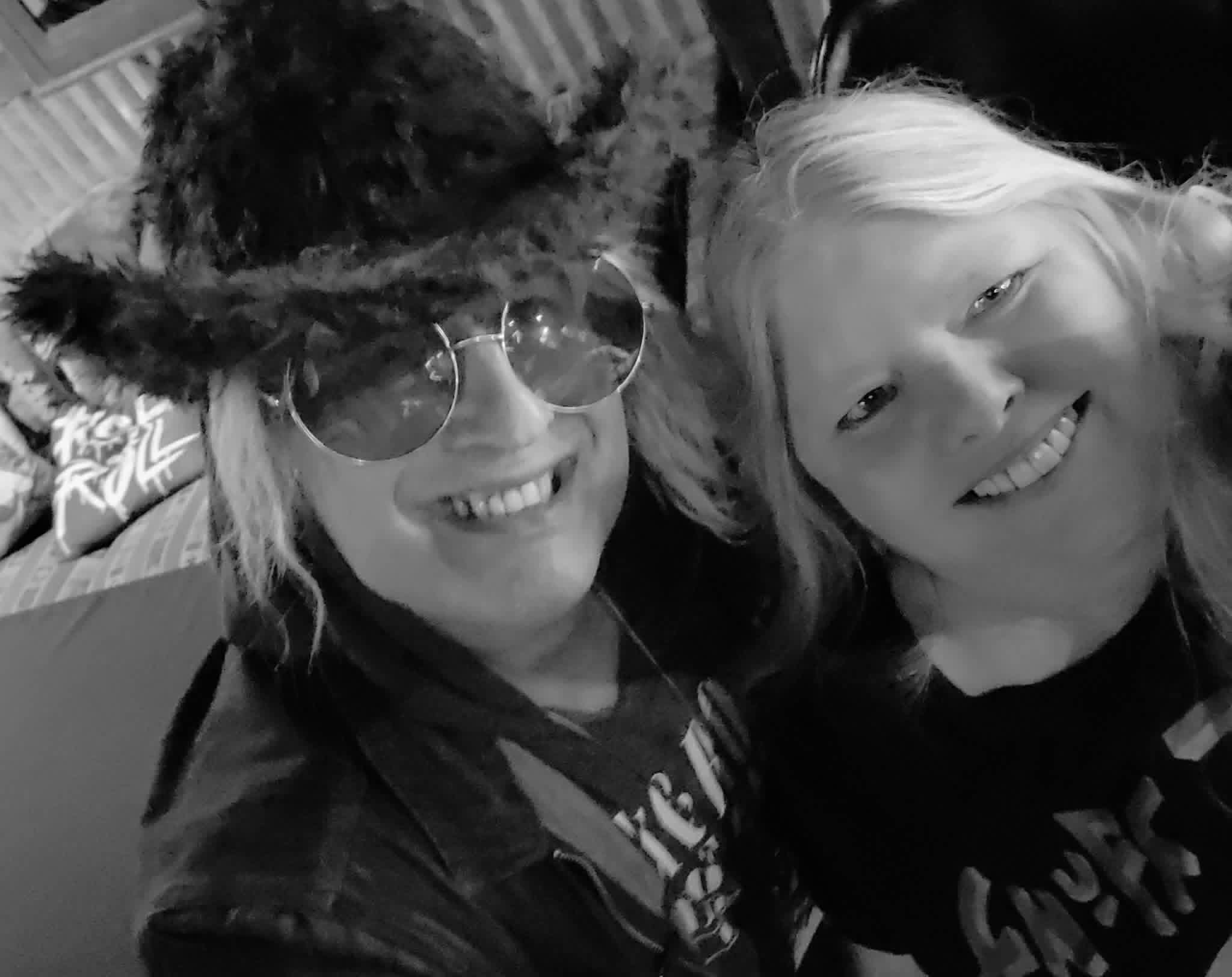 Two smiling people close together, one wearing glasses and a furry hat, the other with long hair, in black and white.
