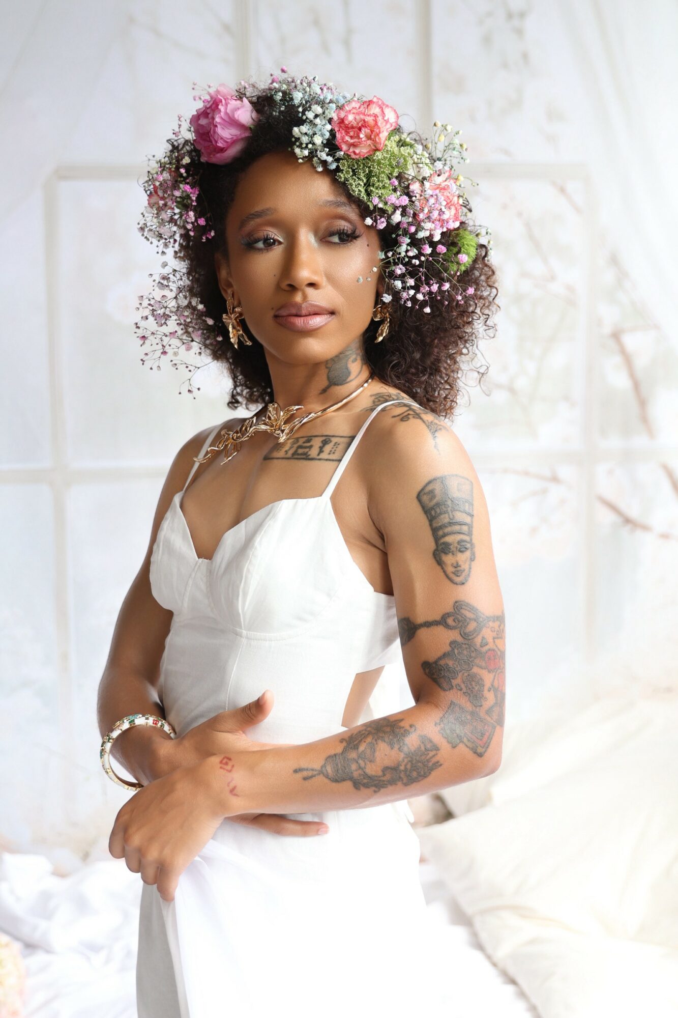 Woman with floral crown, wearing white dress, standing indoors with light background.