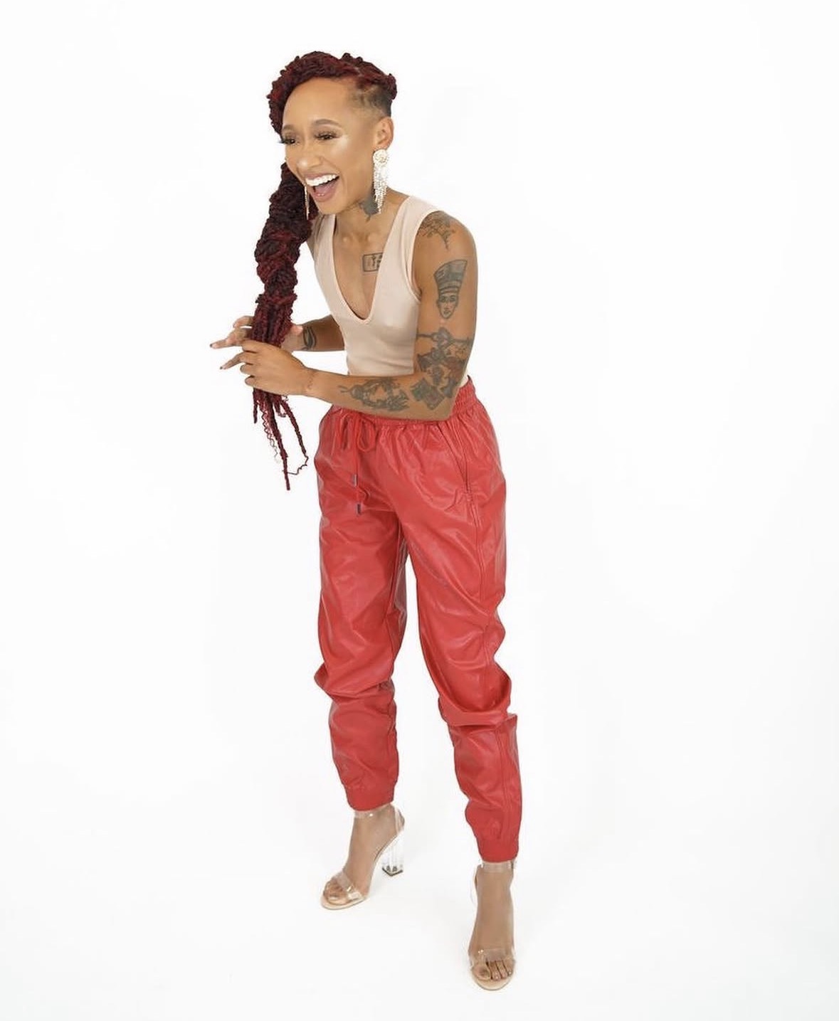 Woman with tattoos laughing, wearing a white top and red pants, standing barefoot against a white background.