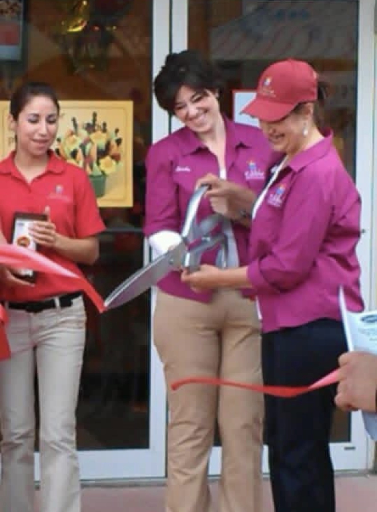 Three women in pink and red uniforms stand outside a store, one holding a red ribbon, celebrating an event.
