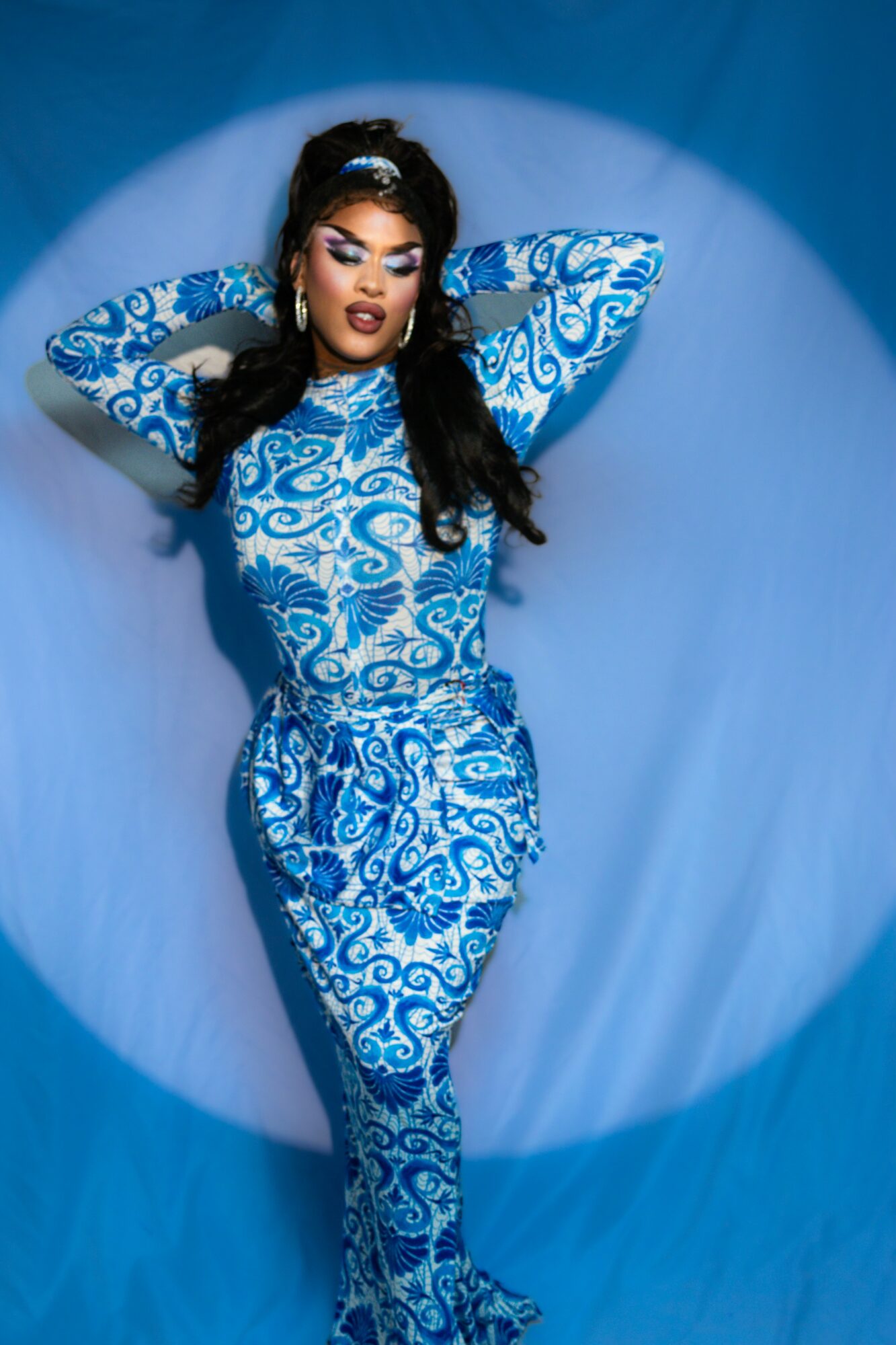 Woman in a blue patterned dress lying on a circular surface with hands behind head, eyes closed, surrounded by blue background.