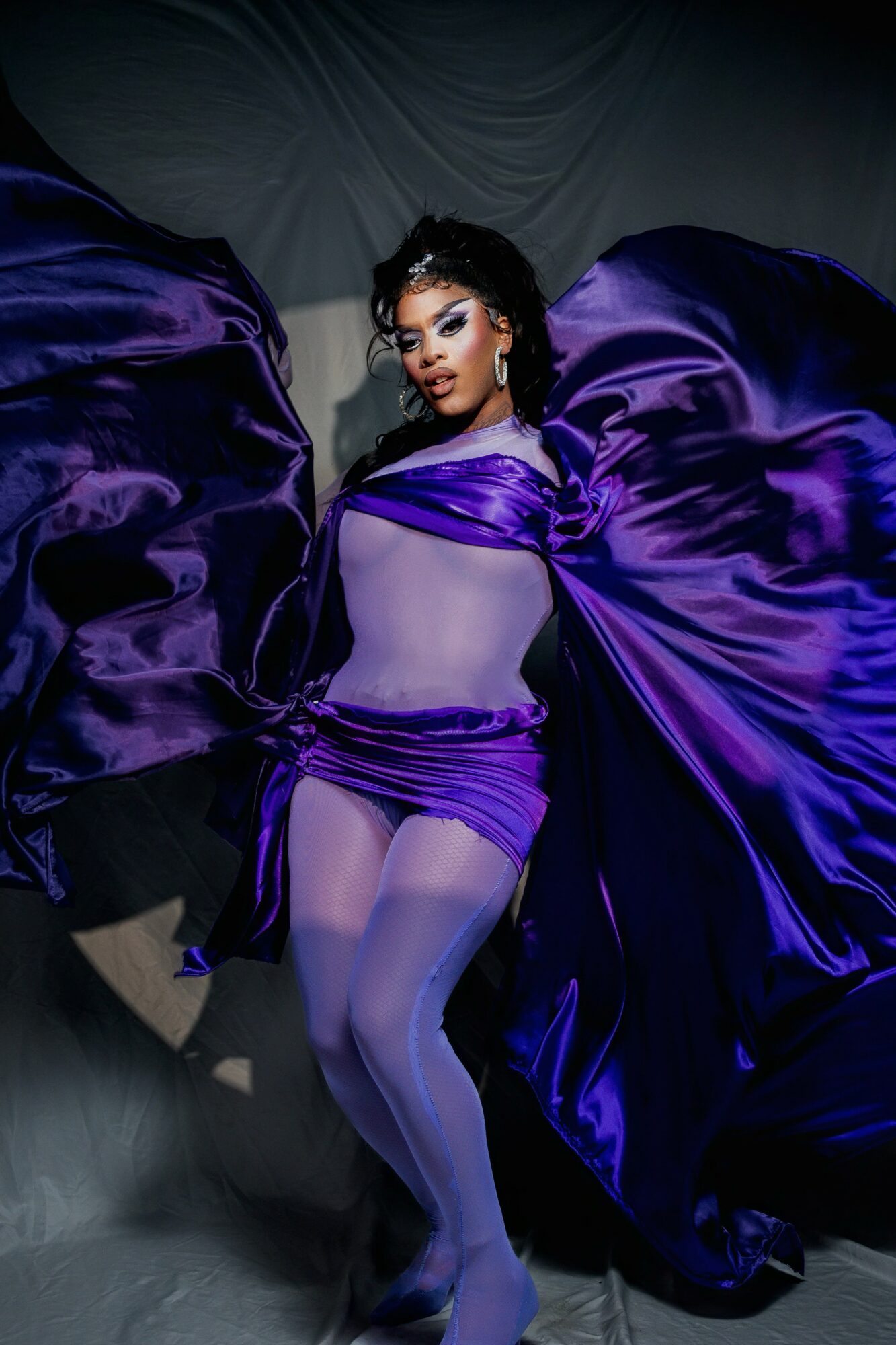 Woman in purple satin outfit with flowing fabric, posing against a dark background.