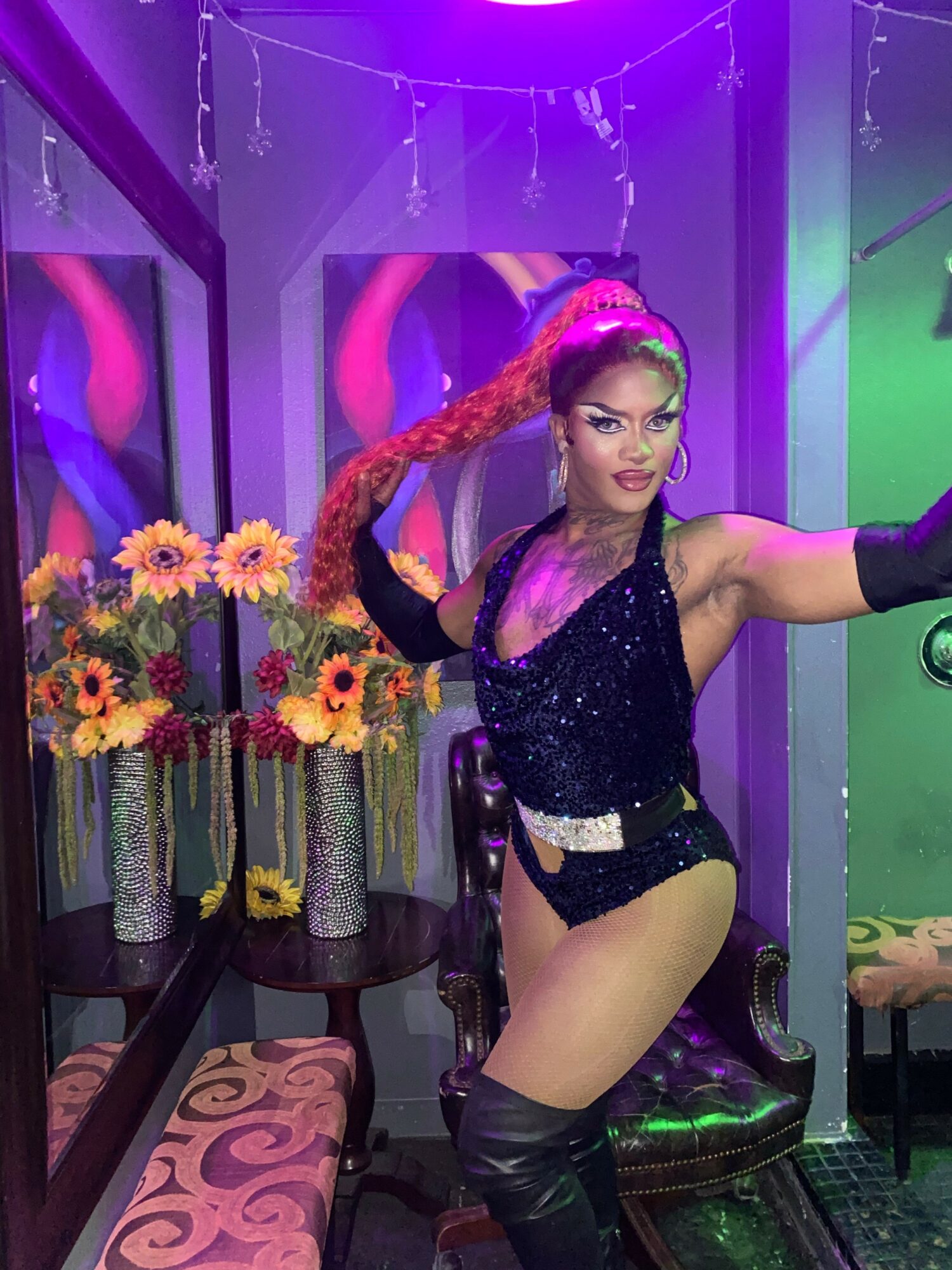 Performer with long red hair, wearing a sparkly black outfit, poses in a colorful room with flowers and purple lighting.
