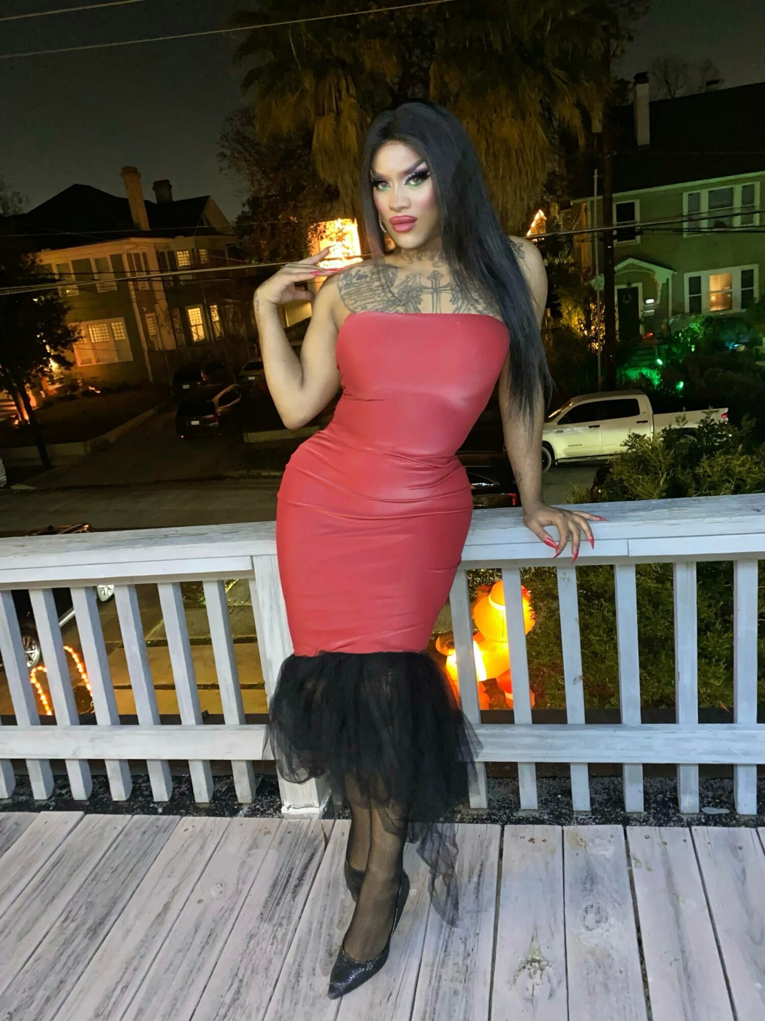 Woman in a red dress with black tulle at the hem, standing on a balcony at night, holding a drink.