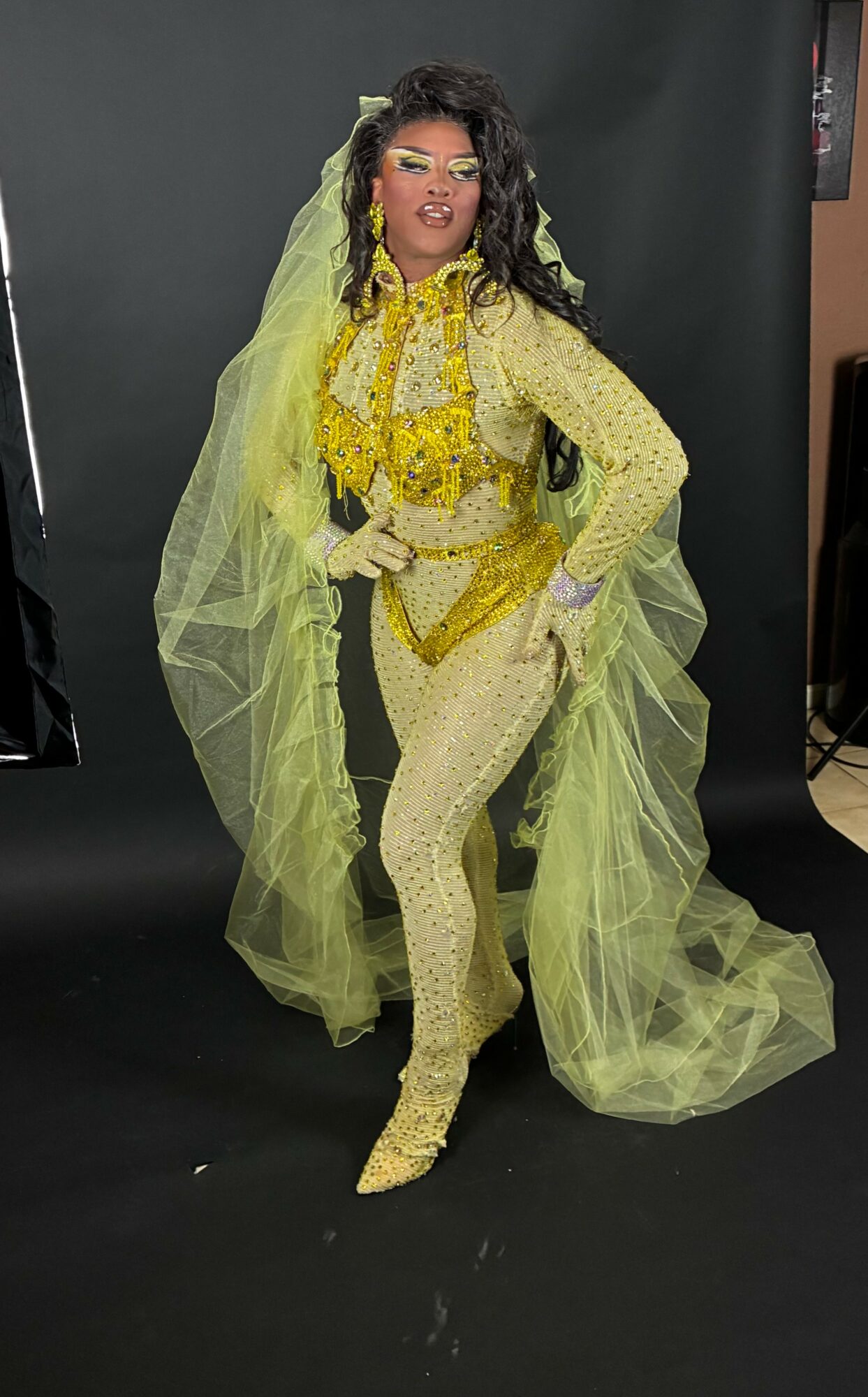 Person in yellow sequined outfit with long veil, standing against a black background, posing confidently with hands on hips.