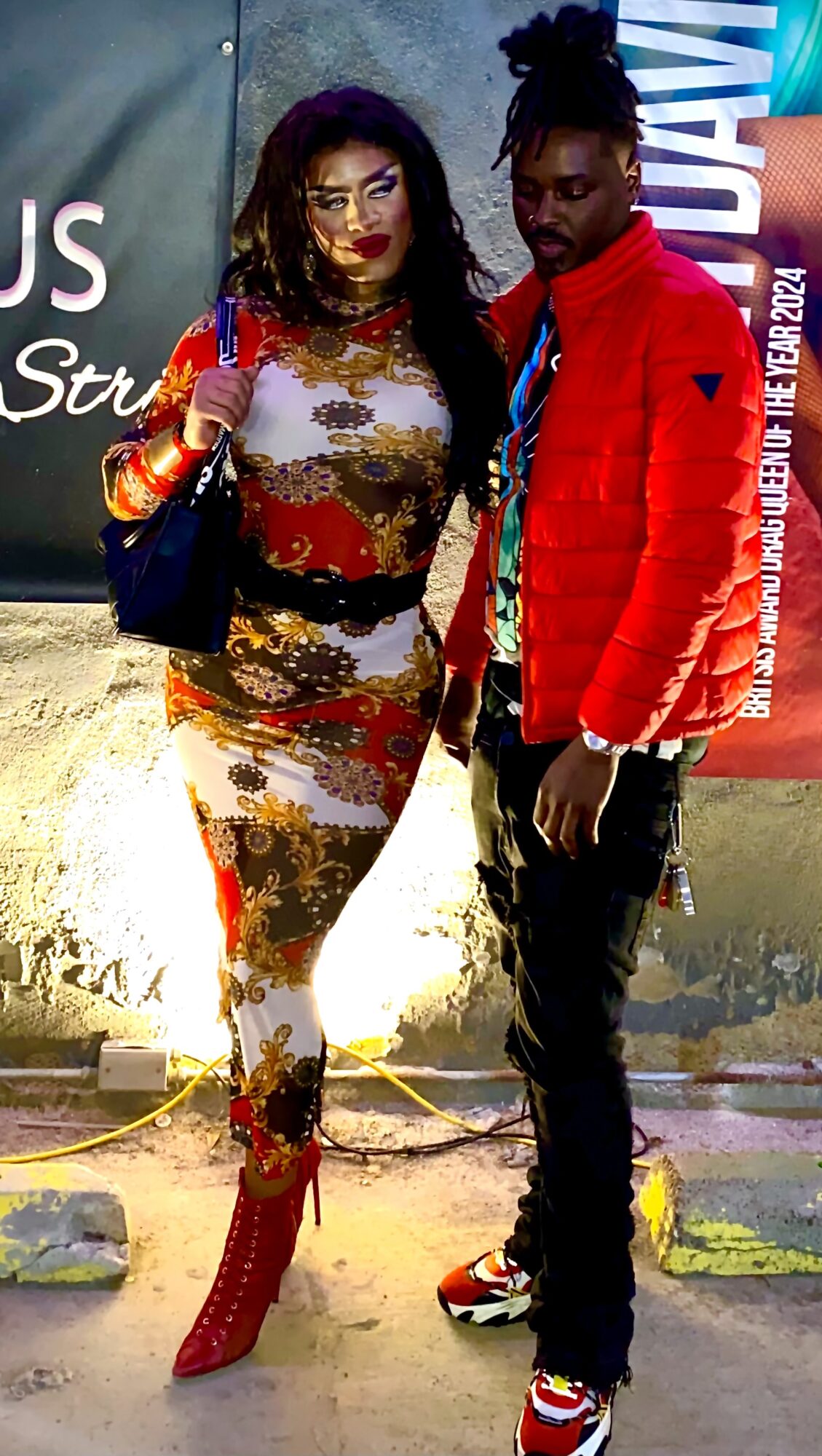 Two people standing outdoors, one wearing a colorful patterned dress and red boots, the other in a red jacket and black pants.